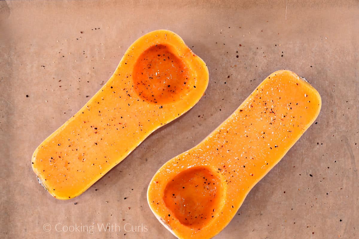 Two butternut squash halves brushed with olive oil and sprinkled with salt and pepper on a parchment lined baking sheet. 