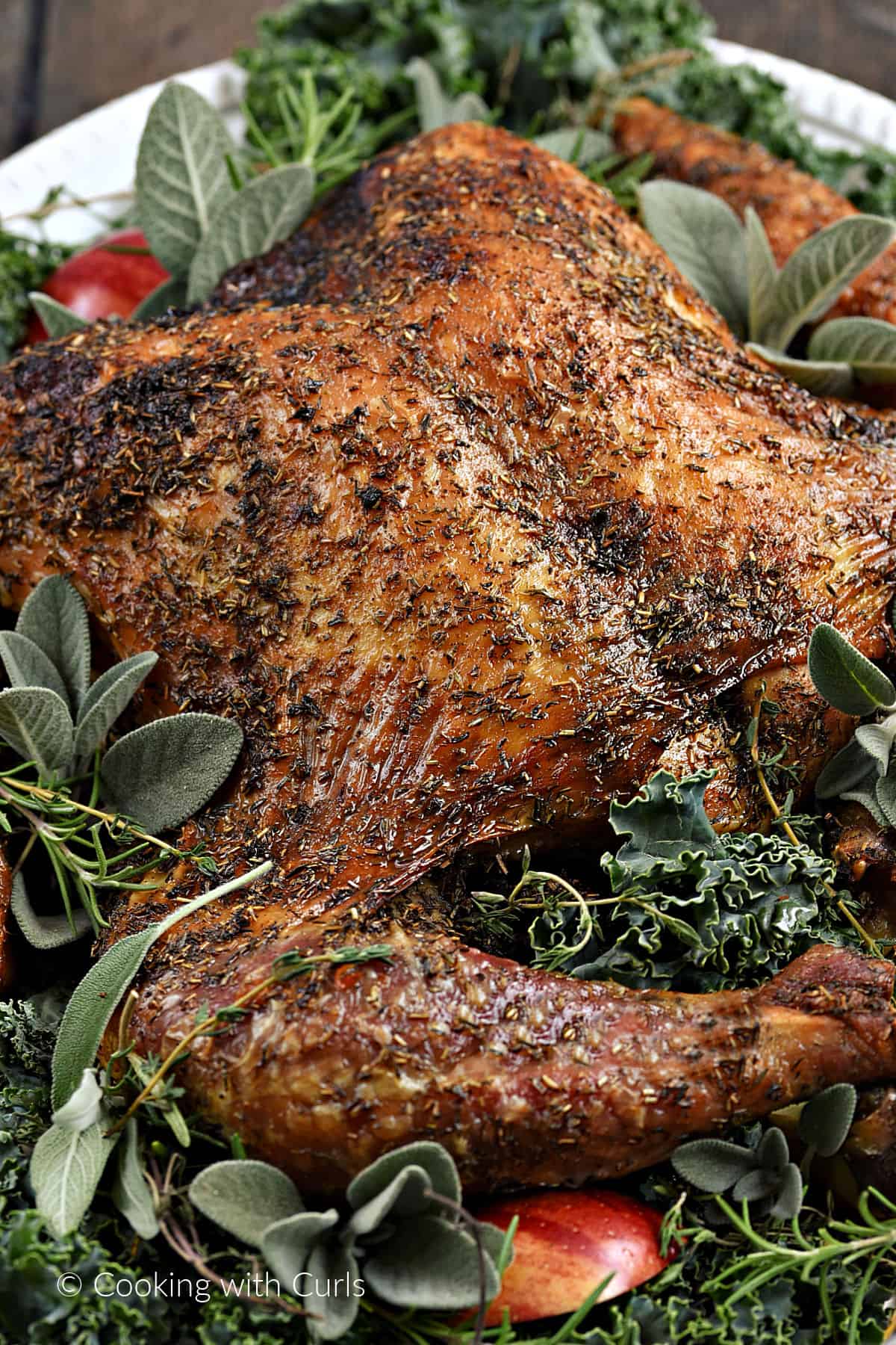 A close-up image of a spatchcock turkey on a plate covered with kale, sliced apples, sage, rosemary and thyme sprigs. 