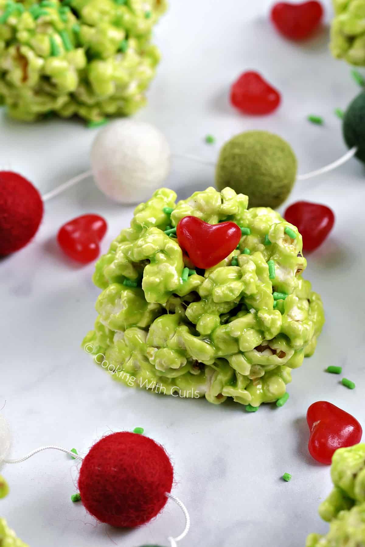 A green marshmallow popcorn ball with a red candy heart and green sprinkles surrounded by sprinkles, hearts, and colorful ball garland. 