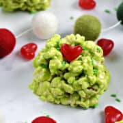 A green marshmallow popcorn ball with a red candy heart and green sprinkles surrounded by sprinkles, hearts, and colorful ball garland.
