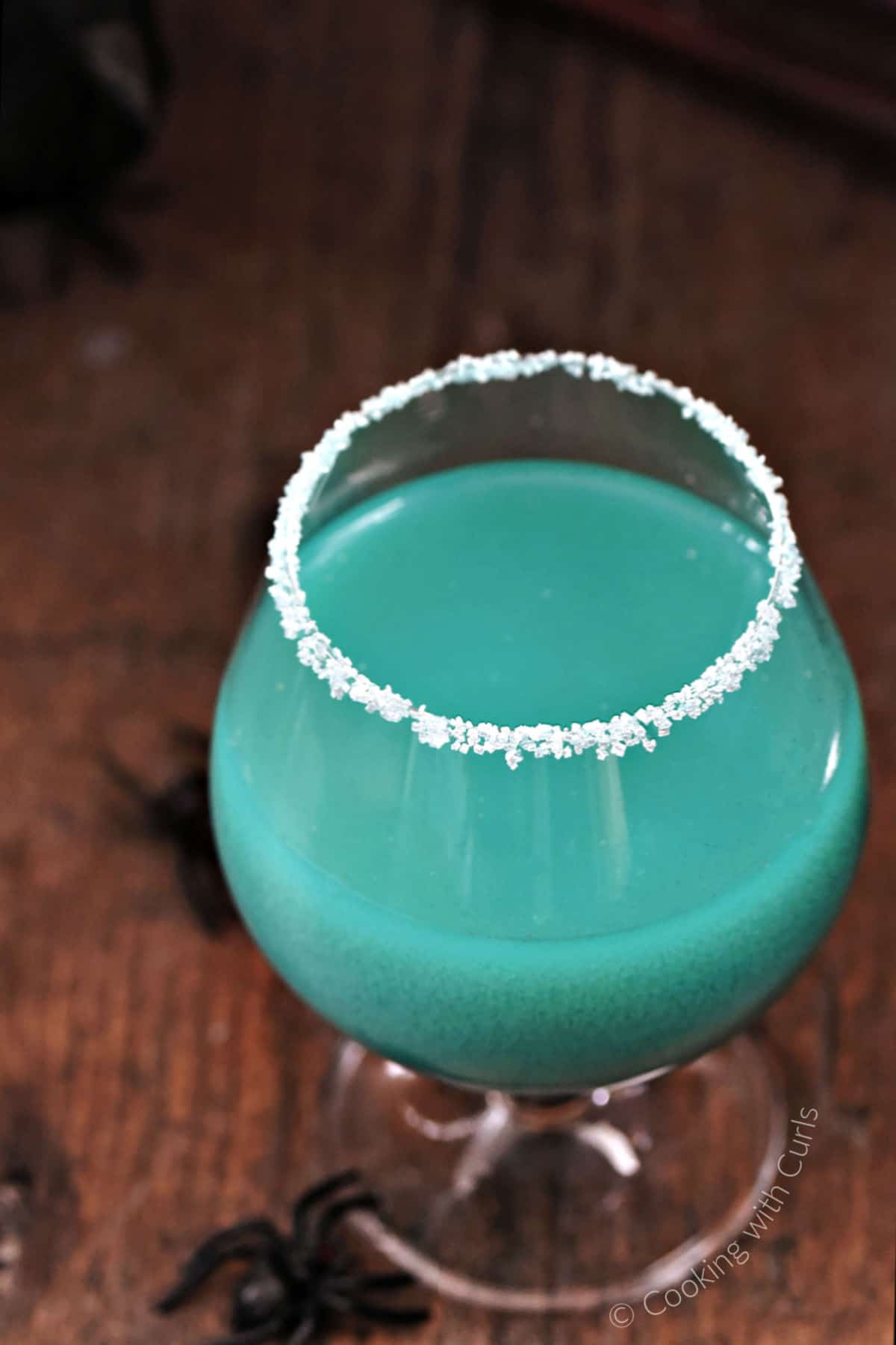 A sugar rimmed goblet with a shimmering teal blue Goblet of Fire Cocktail inside. 