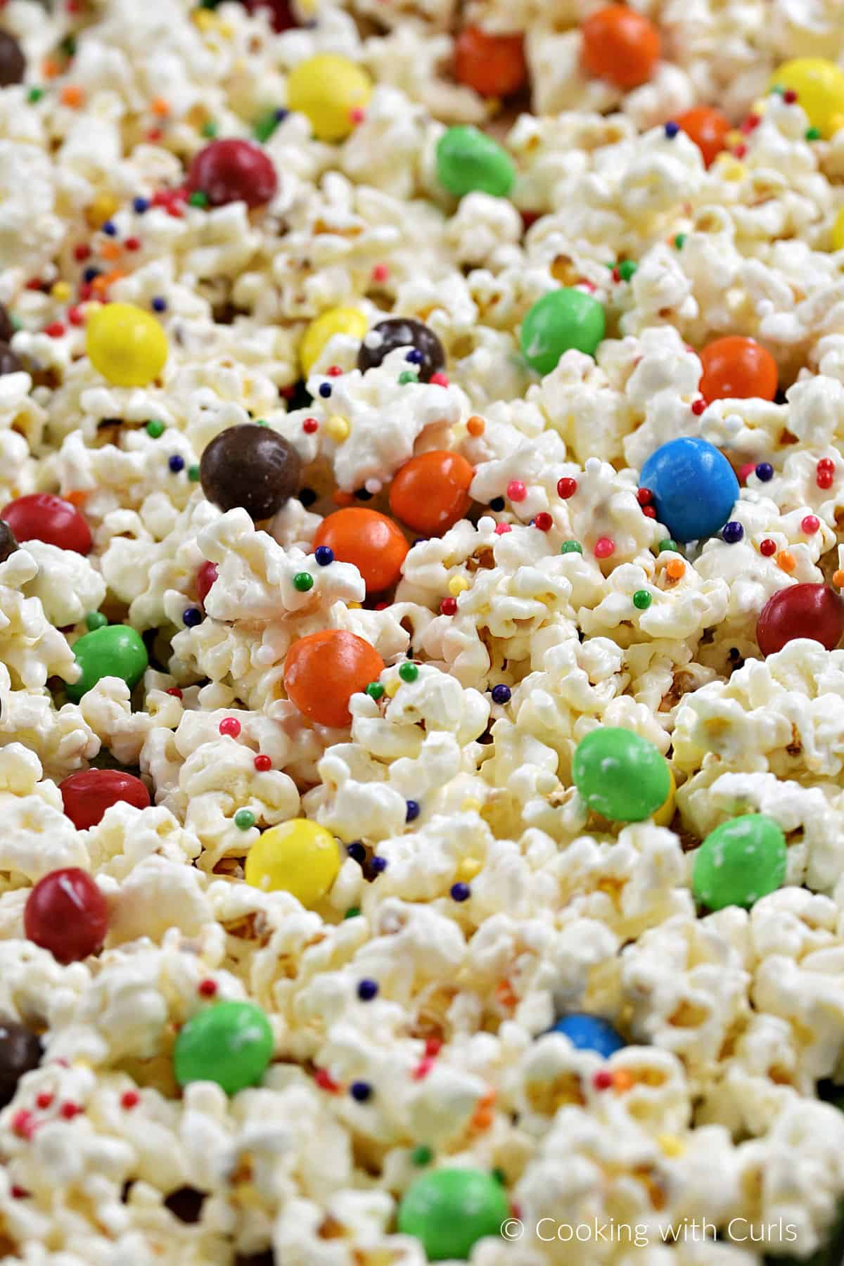 Close-up image of white chocolate popcorn with colored sprinkles and m&ms spread out on a baking sheet. 
