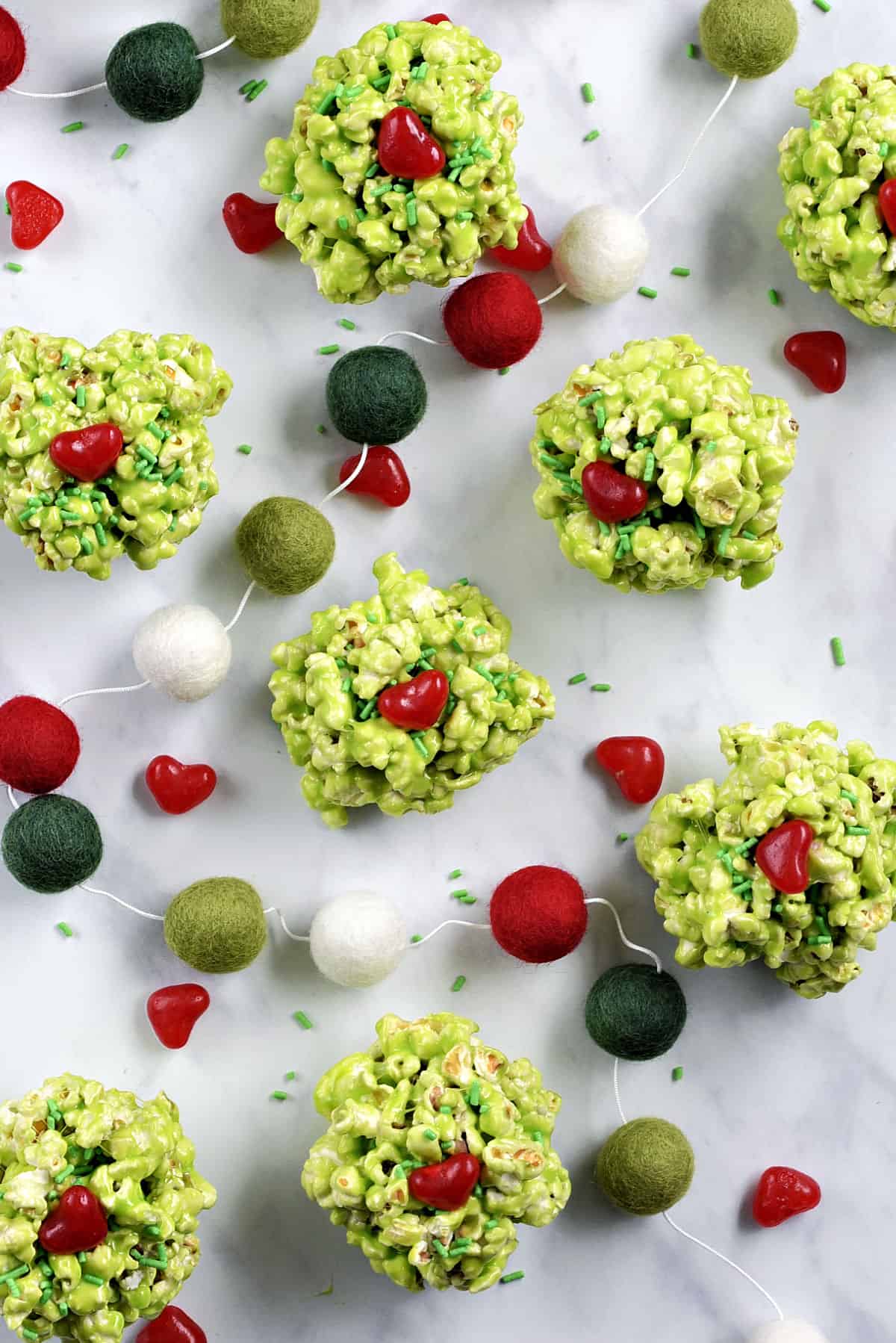 Eight green marshmallow popcorn balls with a red candy heart and green sprinkles and a string of colorful wool balls weaving between them. 