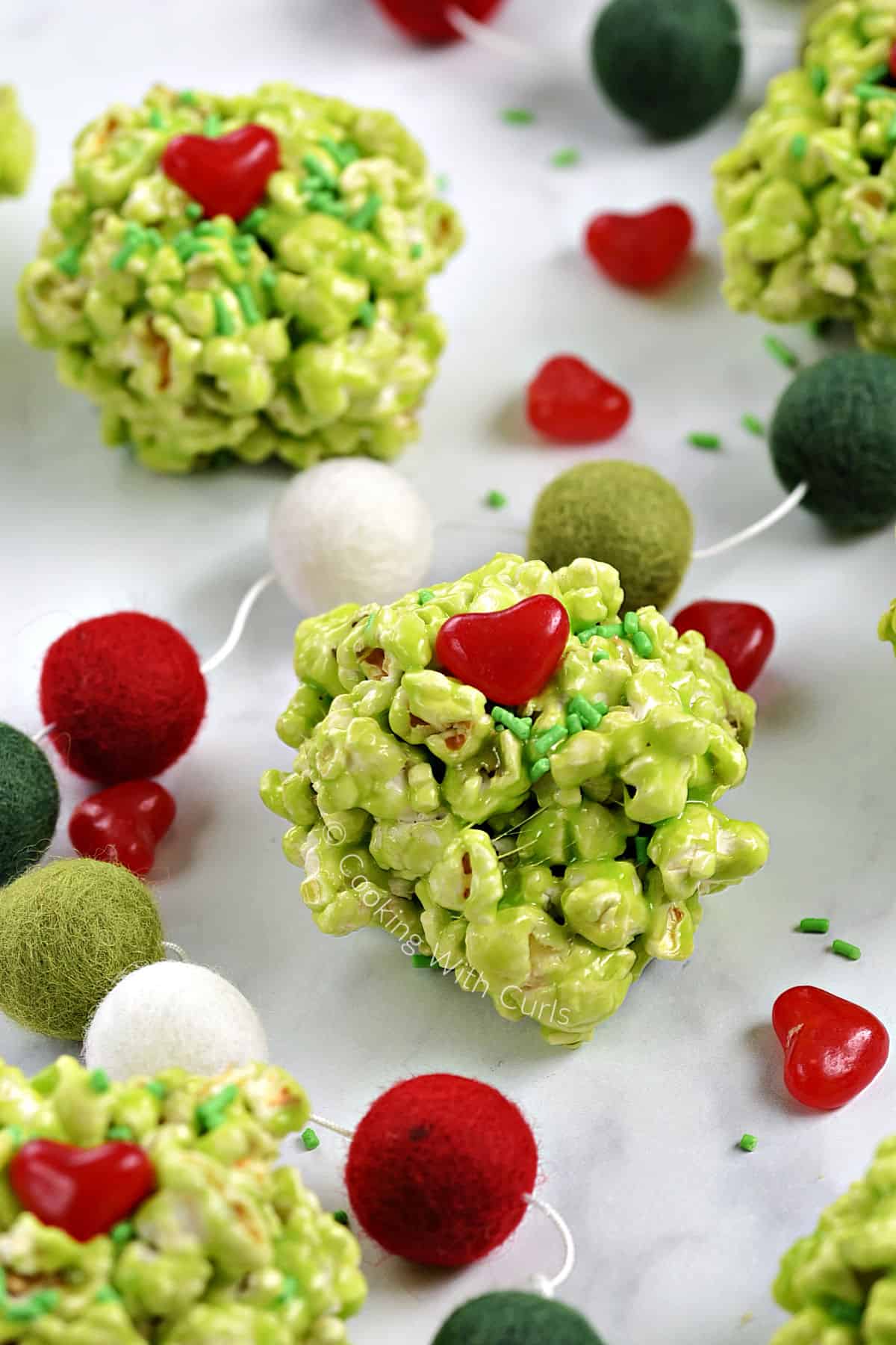Four green marshmallow popcorn balls  with red heart candies and green sprinkles on a board with a string of festive balls. 