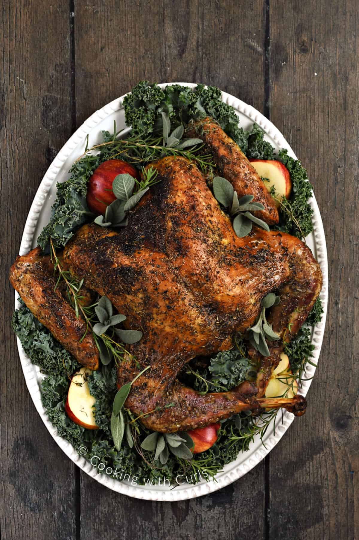 Looking down on a spatchcock turkey on an oval platter covered in kale, sliced apples, sage, rosemary, and thyme sprigs. 