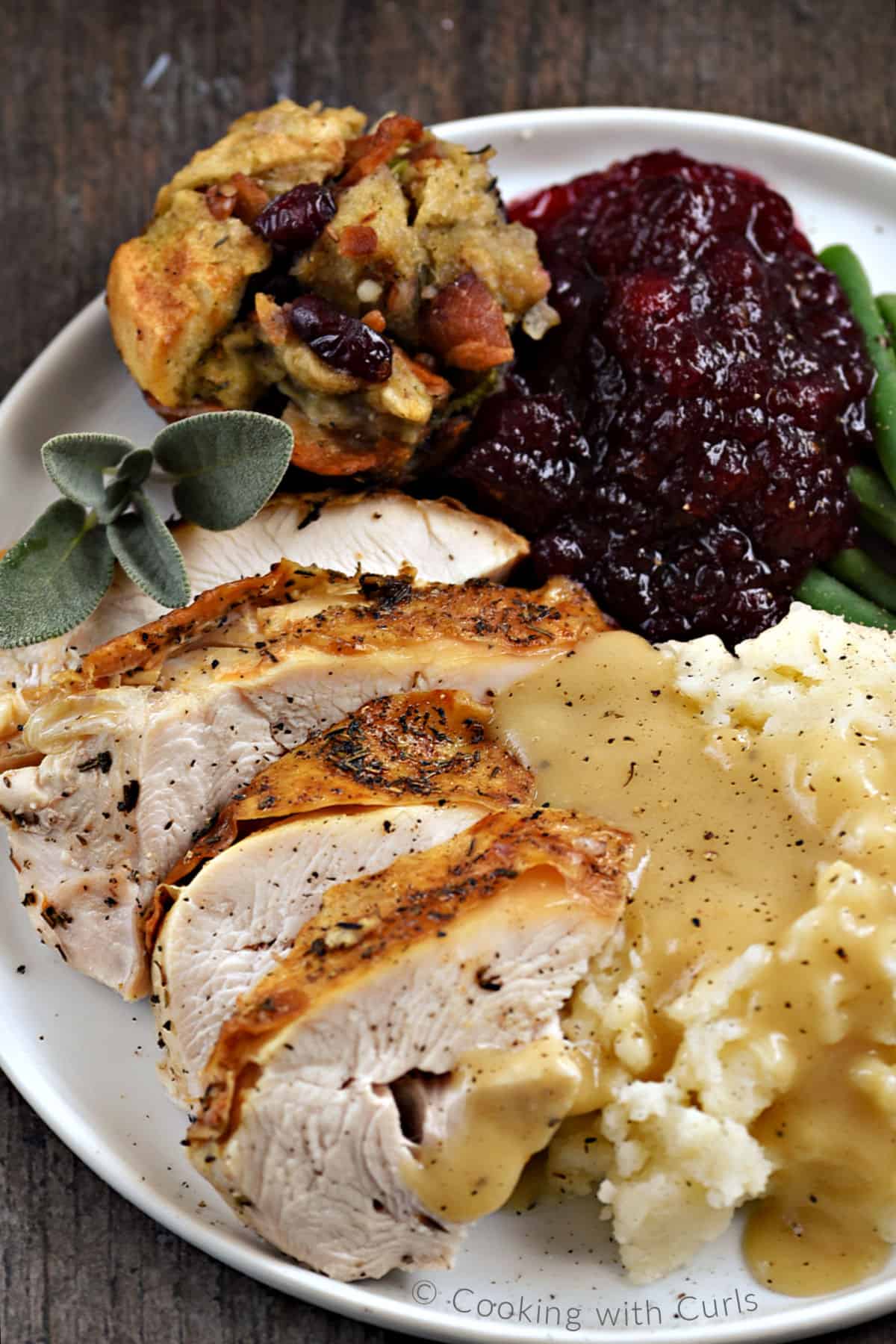Sliced turkey breast, a stuffing muffins, cranberry sauce, green beans, mashed potatoes and gravy on a dinner plate. 