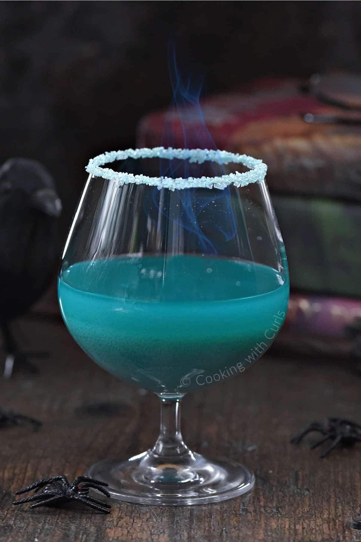 Teal blue Goblet of Fire cocktail in a goblet glass with flames in front of a stack of Harry Potter books.