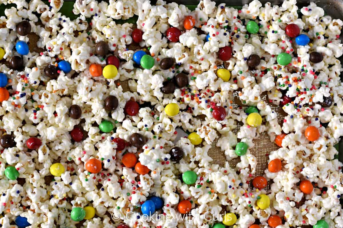 White chocolate covered popcorn, colored sprinkles, and m and m candies spread out on a baking sheet. 