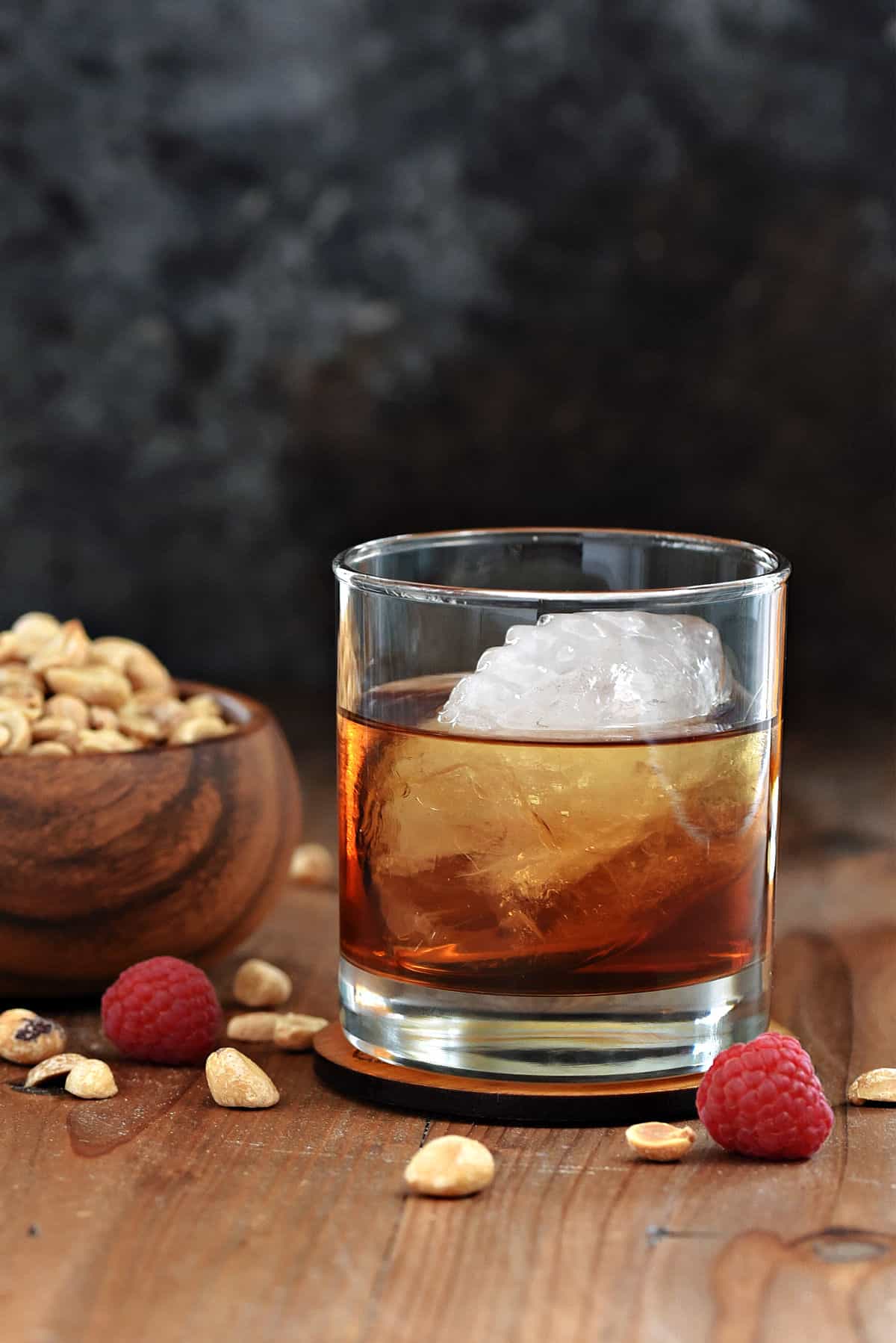 A peanut butter whiskey cocktail in a short glass with a football shaped ice cube and wooden bowl of peanuts in the background. 