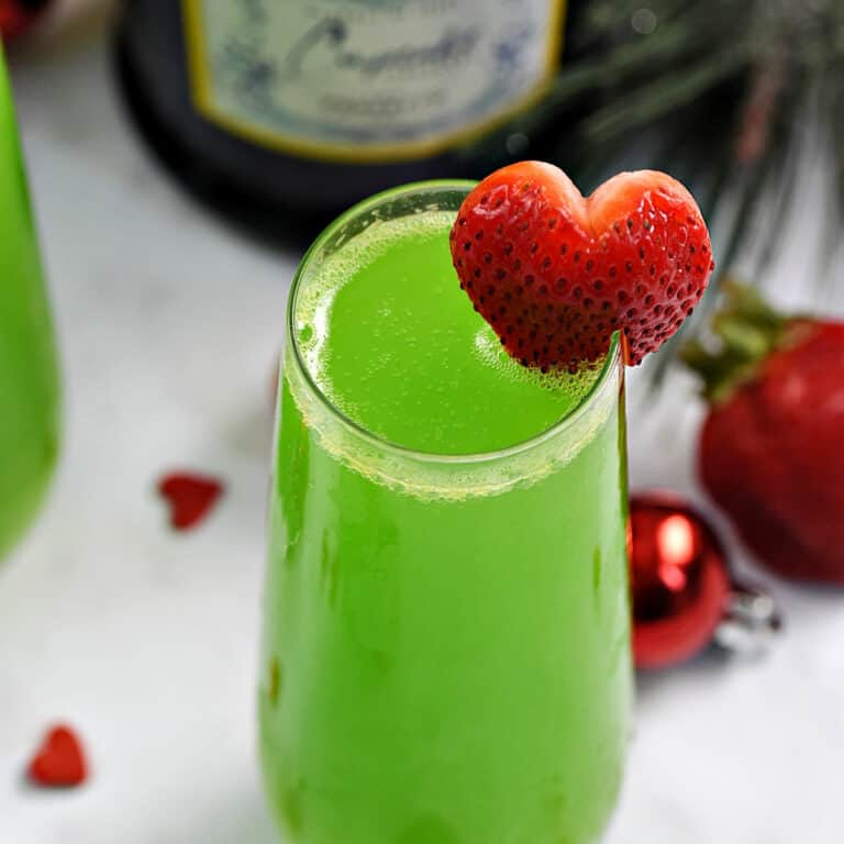 A tall glass of green prosecco cocktail with a strawberry heart garnish and bottle in the background.