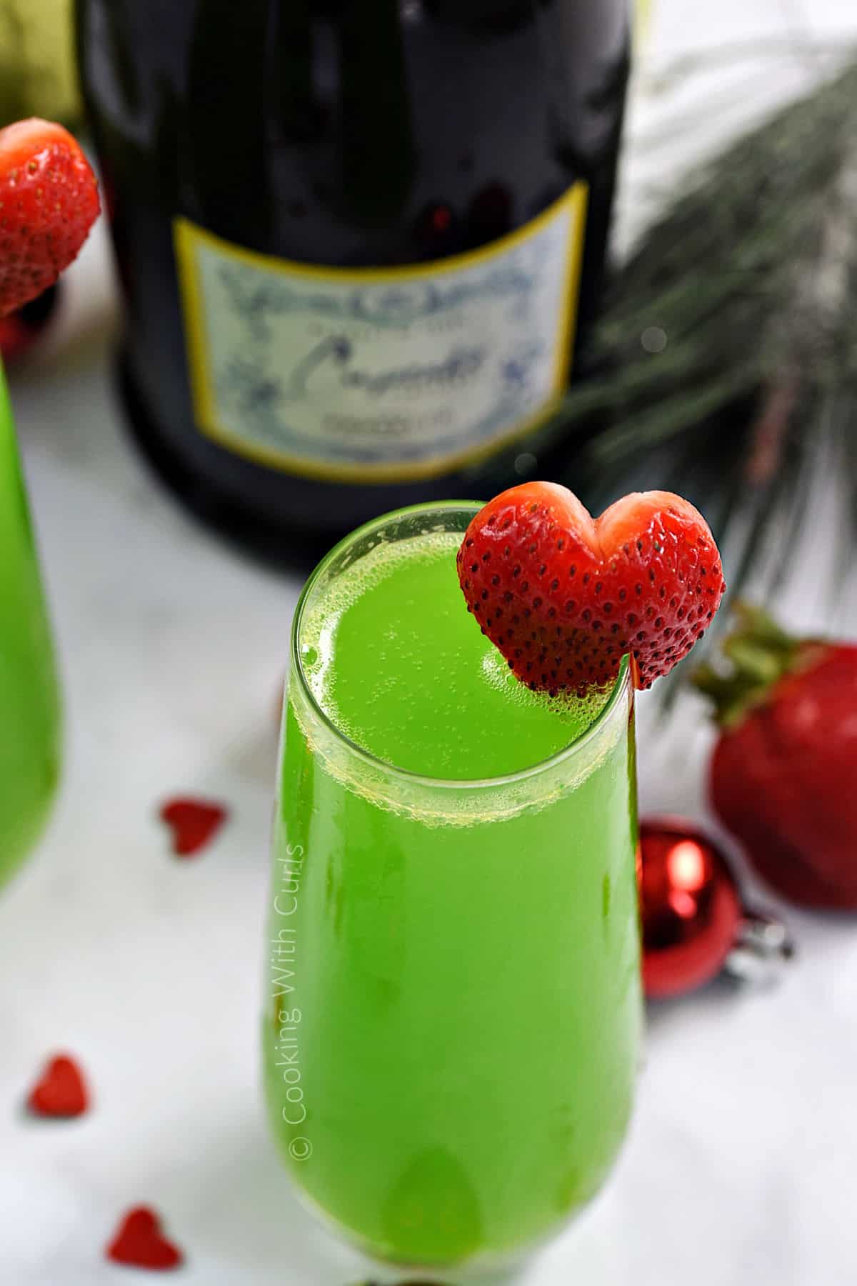 A tall glass of green prosecco cocktail with a strawberry heart garnish and bottle in the background. 