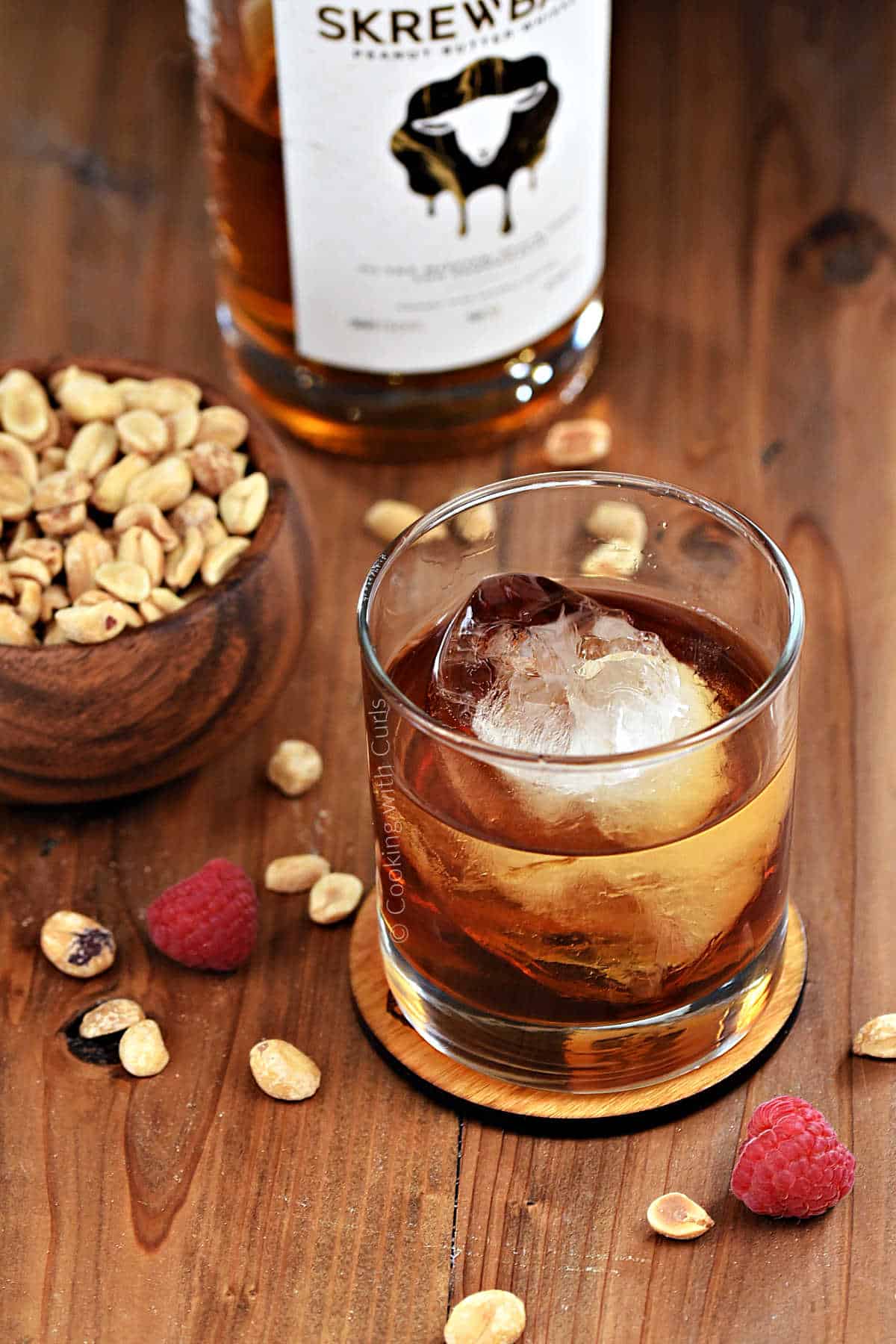 A whiskey cocktail in a short glass with a bottle of peanut butter whiskey, a bowl of peanuts and fresh raspberries in the background. 