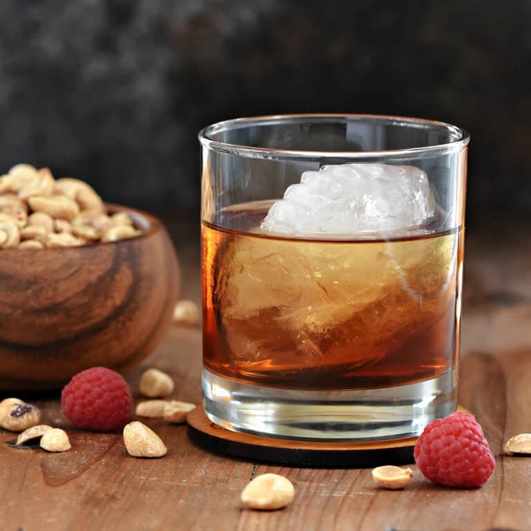 A whiskey cocktail recipe in a short glass with a football shaped ice cube and wooden bowl of peanuts in the background.