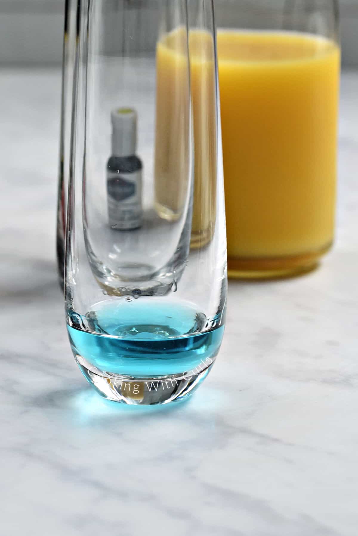 Blue vodka in the bottom of a tall champagne glass with orange juice in the background. 