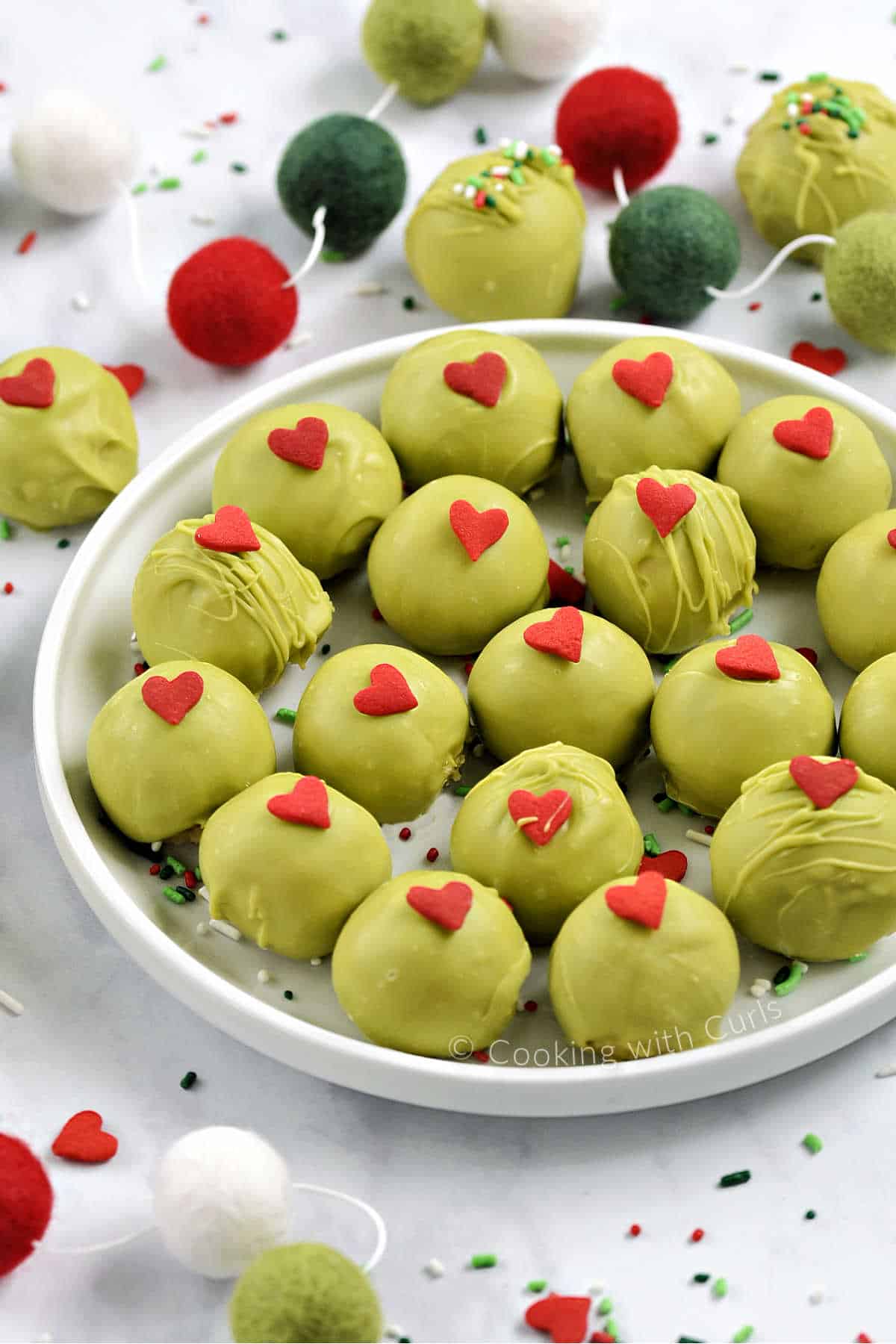 Eighteen green chocolate covered oreo balls topped with red candy hearts on a plate surrounded by ball garland and holiday sprinkles. 