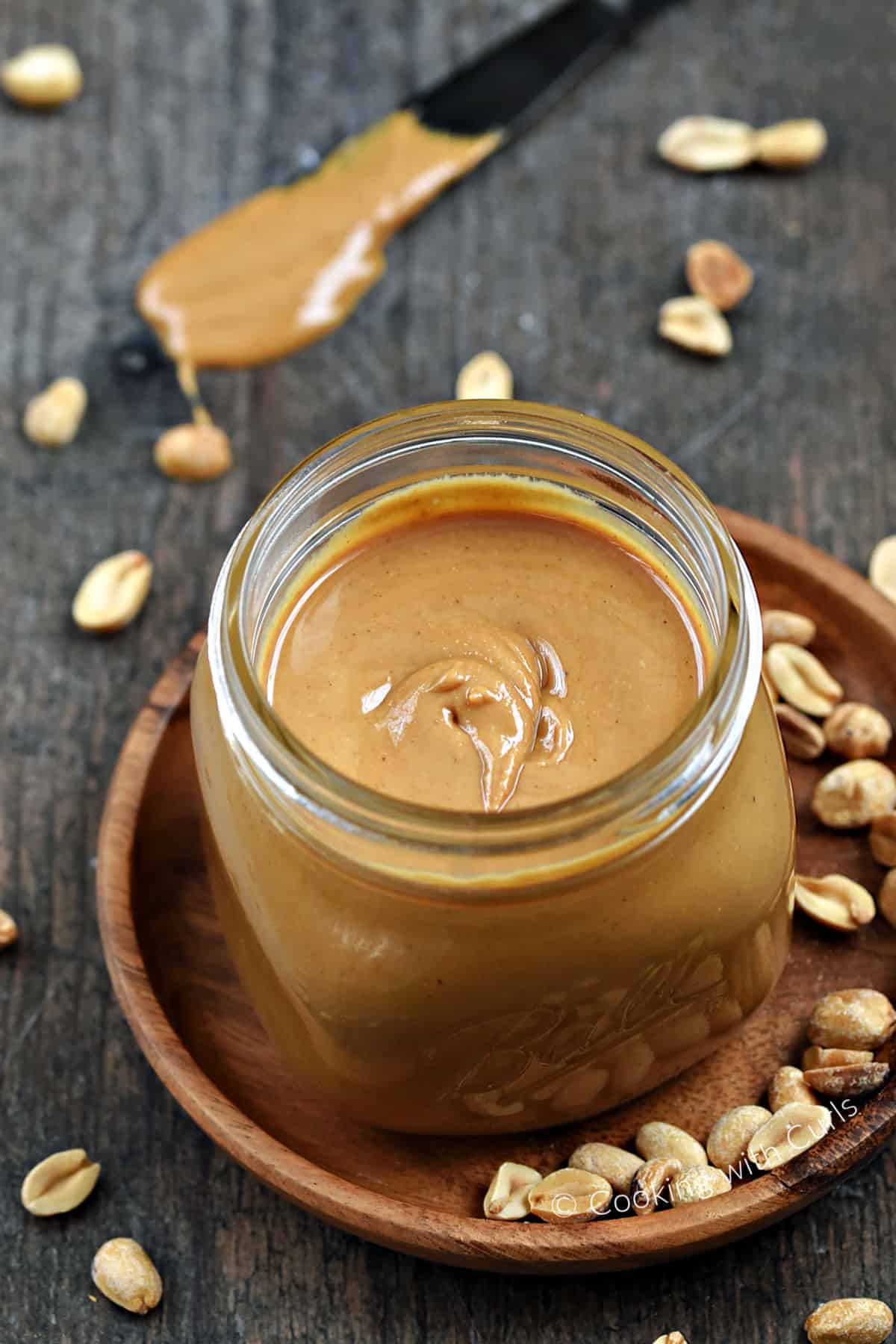 Homemade Peanut Butter in a square jar on a wood plate surrounded by peanuts. 