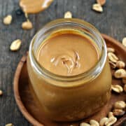 Homemade Peanut Butter in a square jar on a wood plate surrounded by peanuts.