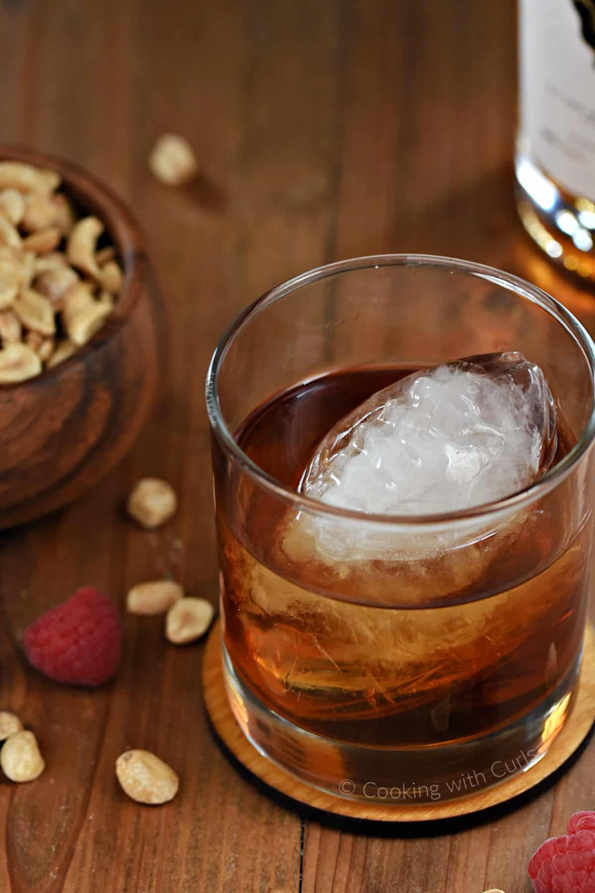 Looking down into a cocktail glass filled with peanut butter whiskey and a football shaped ice cube with a bowl of peanuts in the background. 