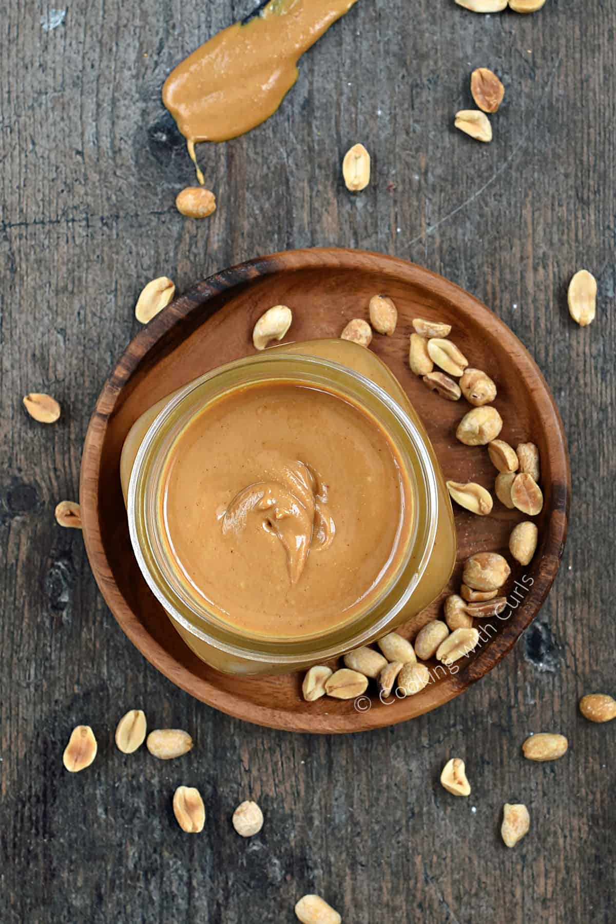 Looking down on a jar of peanut butter on a wood plate surrounded with peanuts and a peanut butter covered knife in the background. 