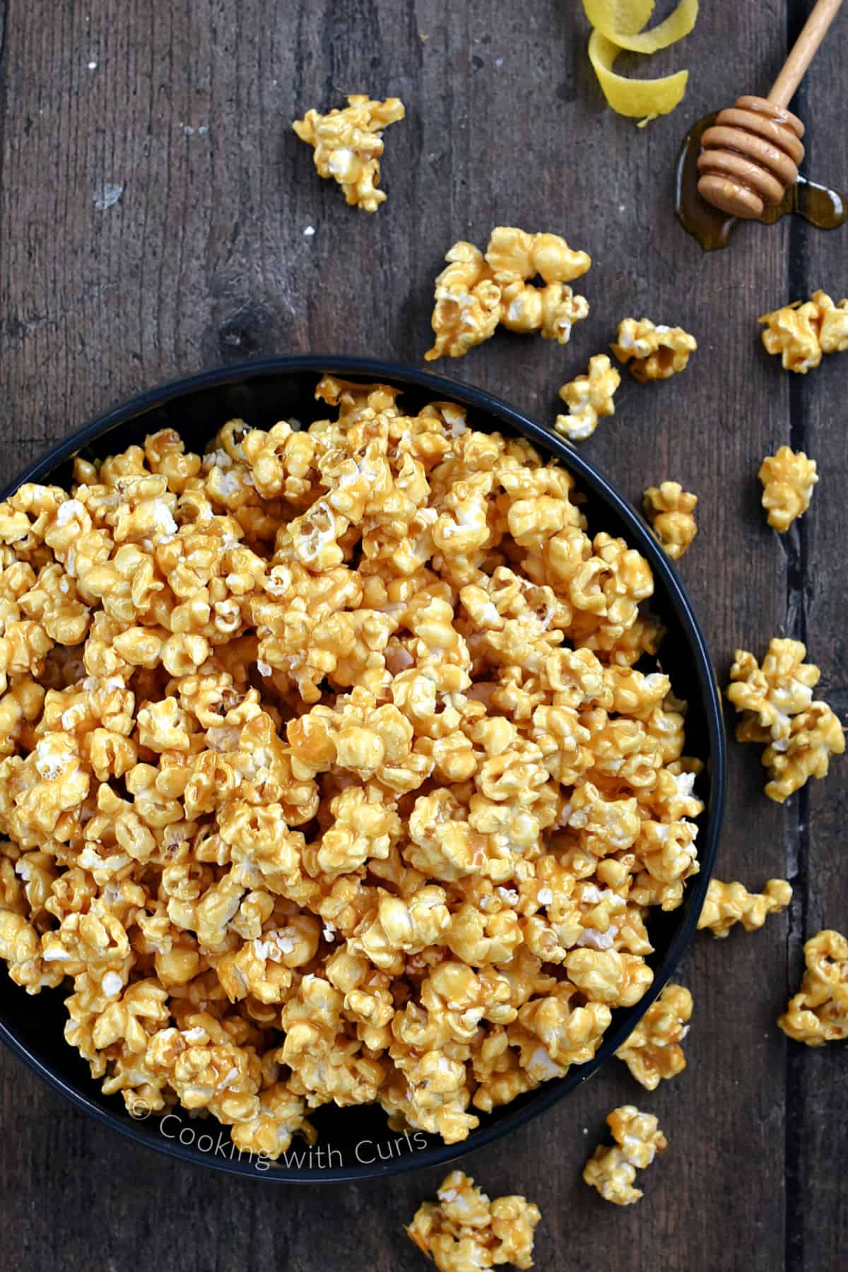 Looking down on a large bowl of caramel popcorn with spilled popcorn, honey and lemon rinds in the background. 