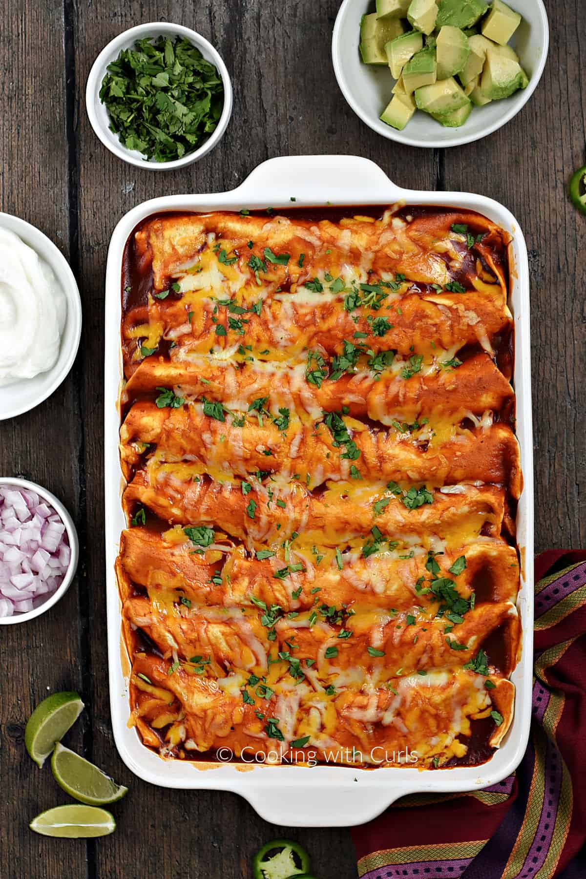 Looking down on a pan of turkey enchiladas with bowls of sour cream, red onion, cilantro, and avocado chunks around the edge. 