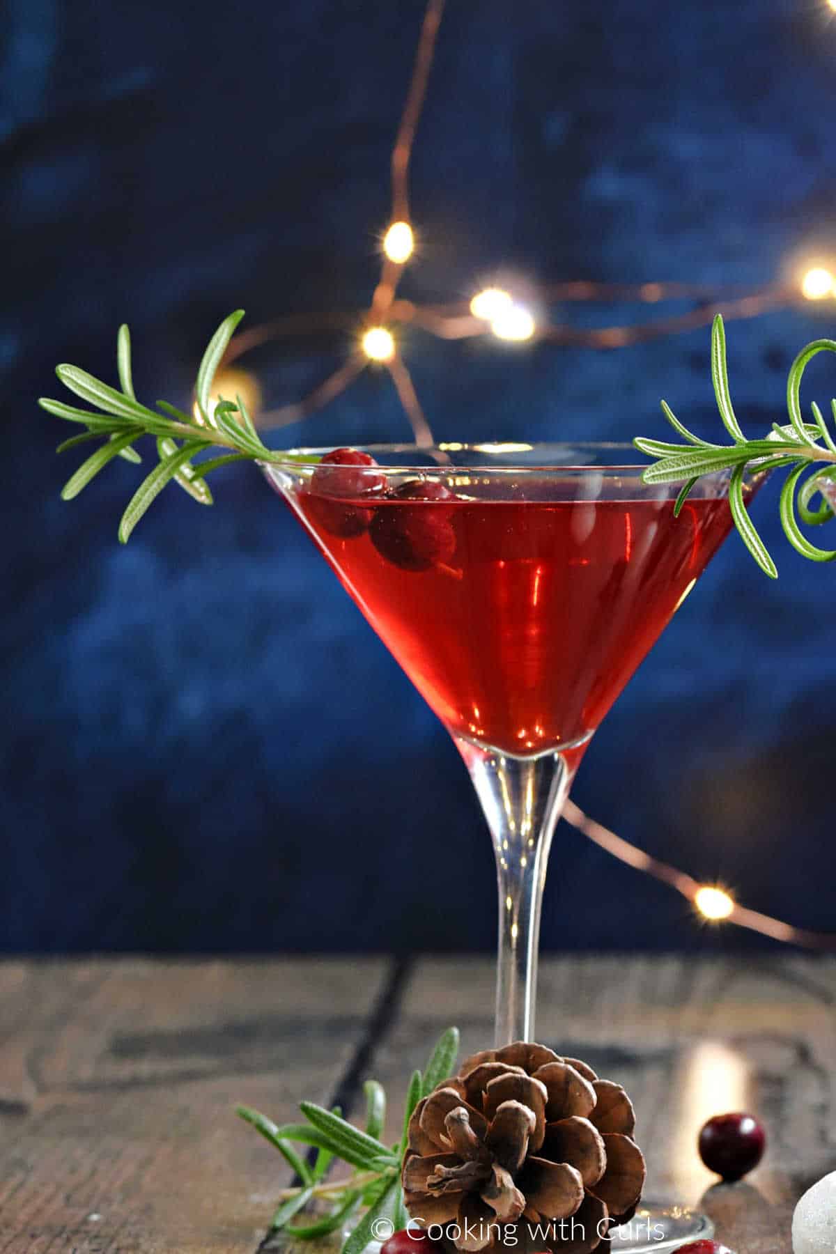 Bright red cocktail in a martini glass garnished with rosemary and cranberries. 