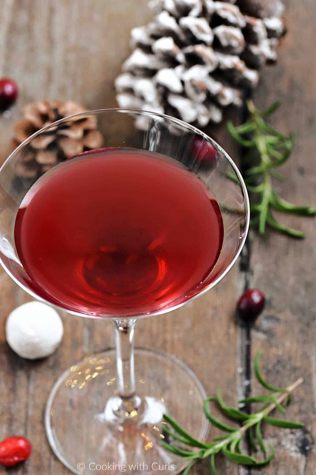 Cranberry juice, vanilla vodka, and seltzer in a martini glass surrounded by rosemary, cranberries, and pine cones. 