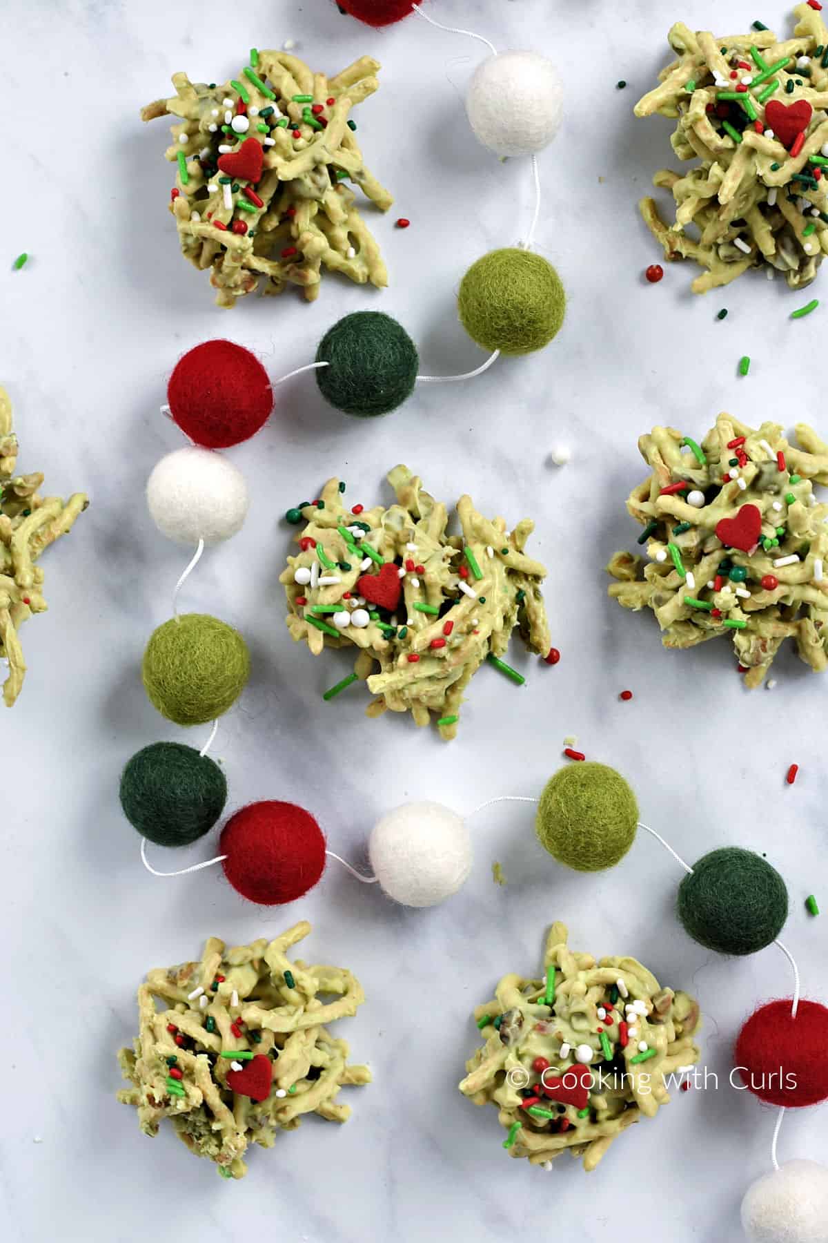 Looking down on seven green chocolate Grinch Haystack Cookies with red hearts and decorative holiday sprinkles  with garland twisted through the center. 