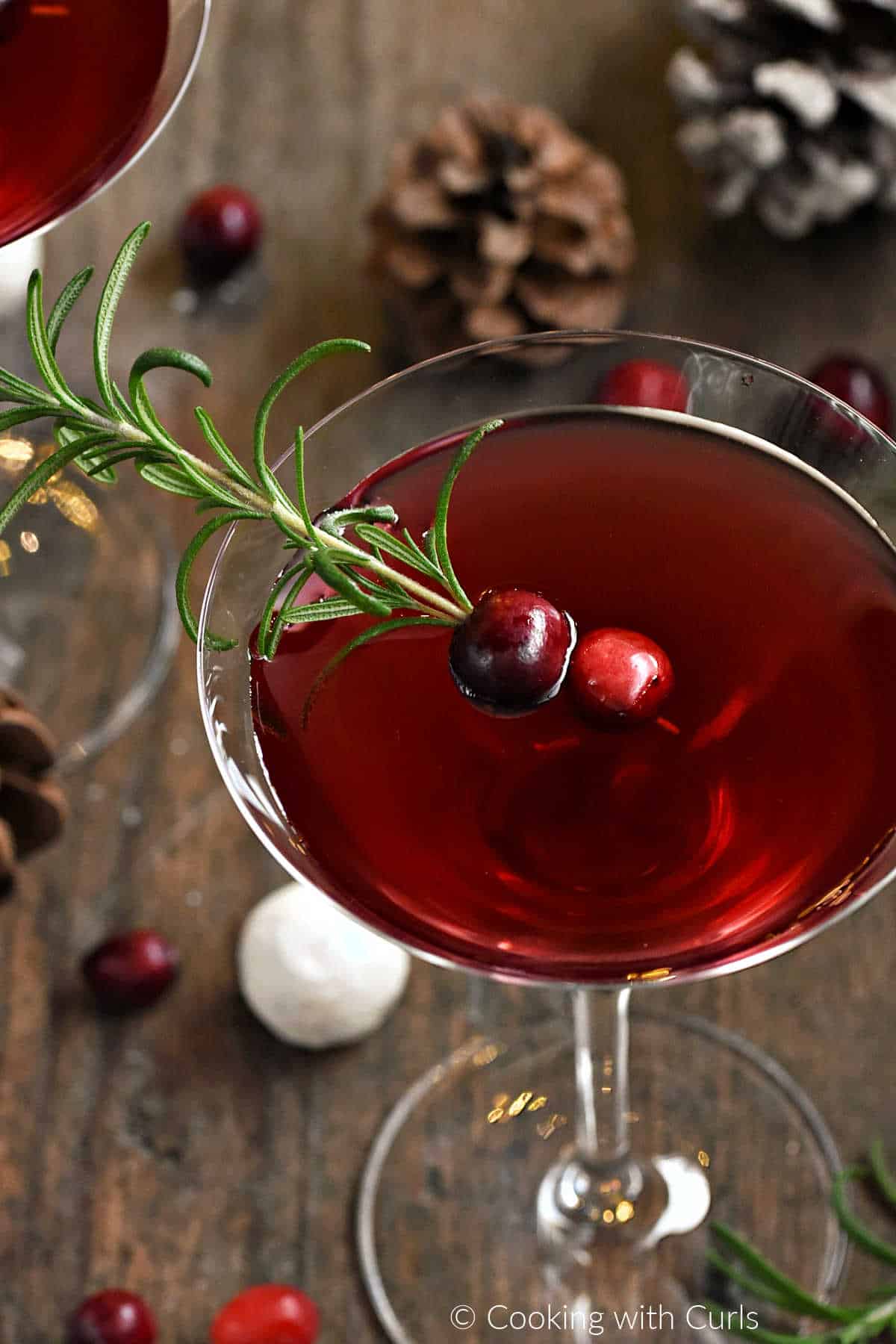 Red Christmas Martini recipe garnished with fresh rosemary sprig and two cranberries surrounded by pine cones. 