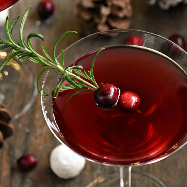 Red Christmas Martini recipe garnished with fresh rosemary sprig and two cranberries surrounded by pine cones.