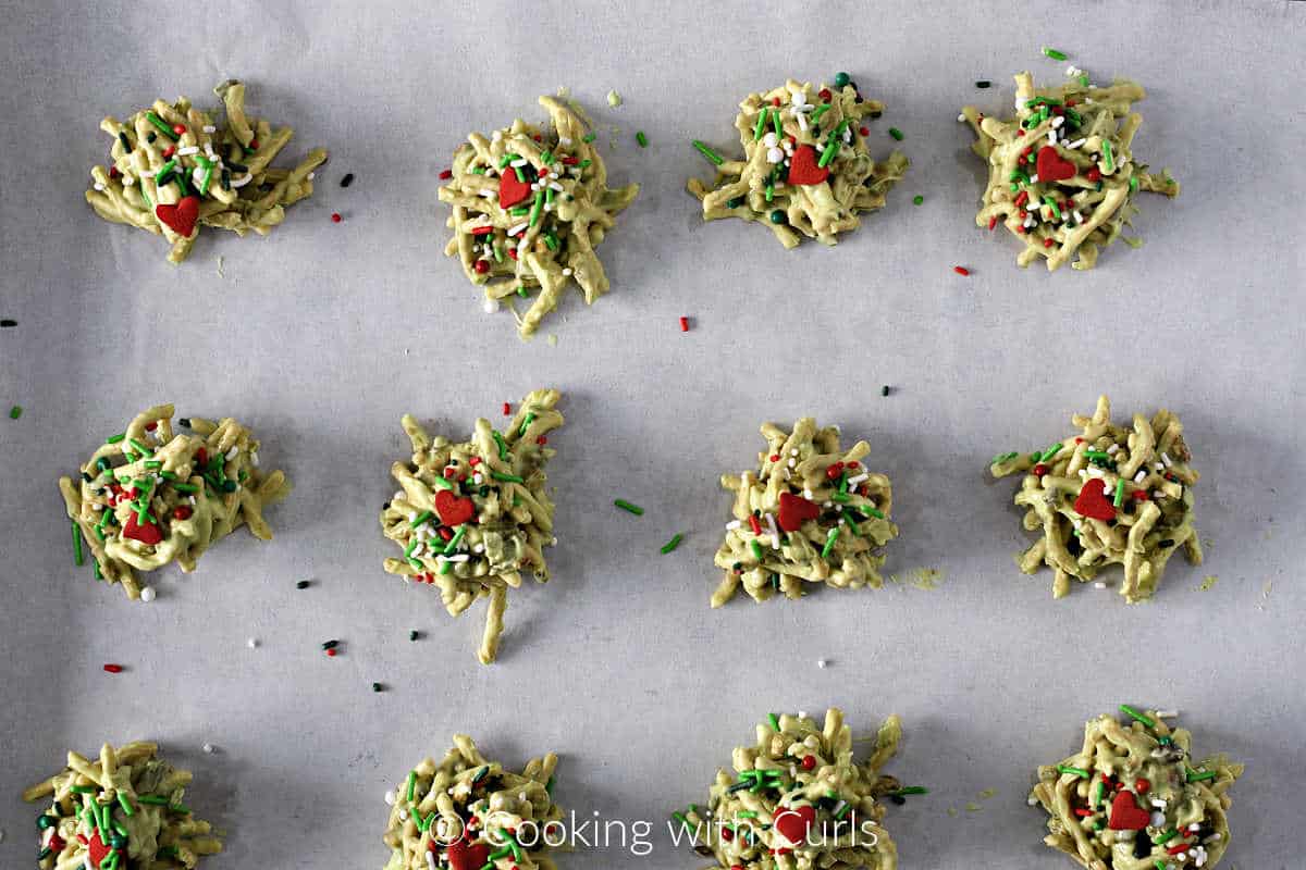 Twelve green chocolate covered chow mein cookies with sprinkles and red hearts on a sheet of parchment paper. 
