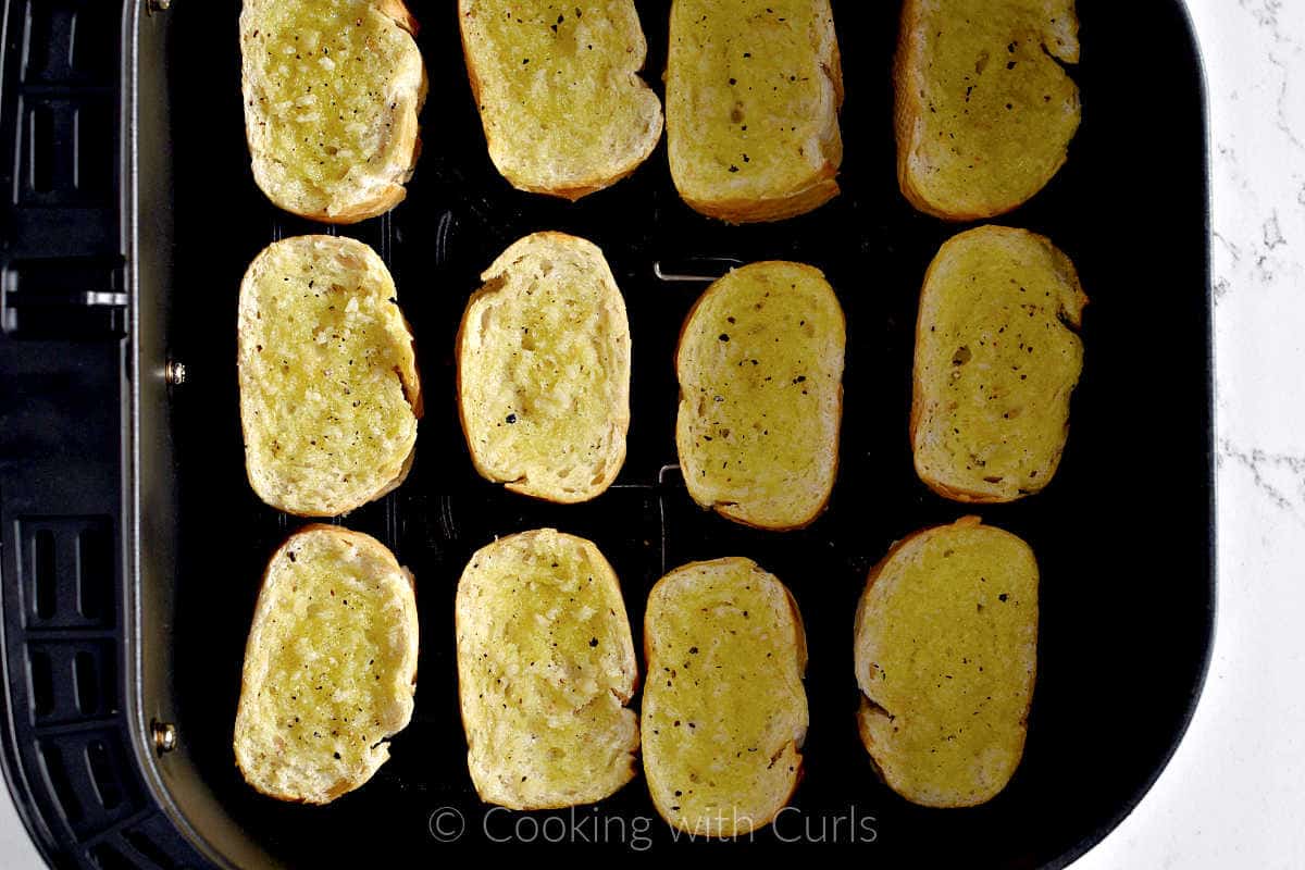 Twelve slices of oil topped baguettes in an air fryer basket. 