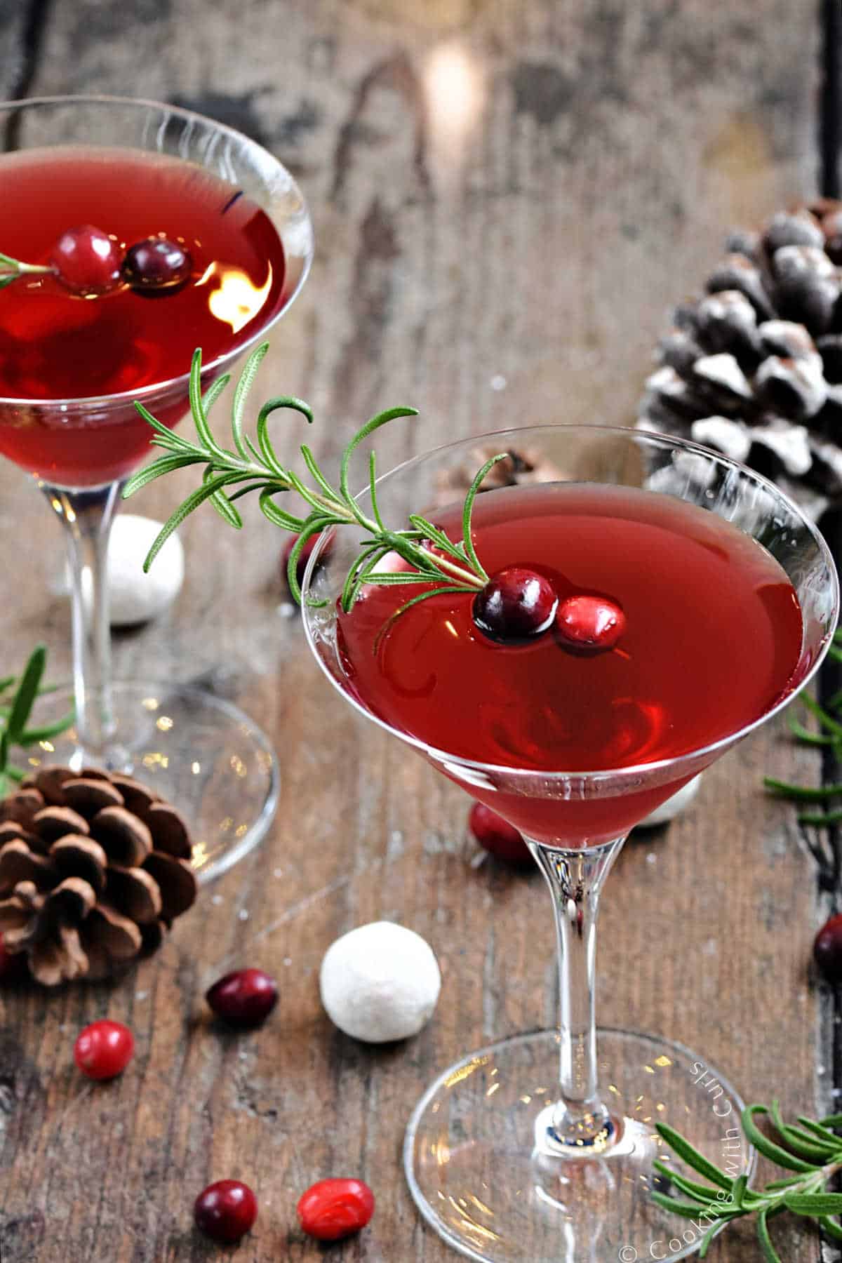 Two, red Christmas Martinis garnished with rosemary sprigs and cranberries surrounded by pine cones. 