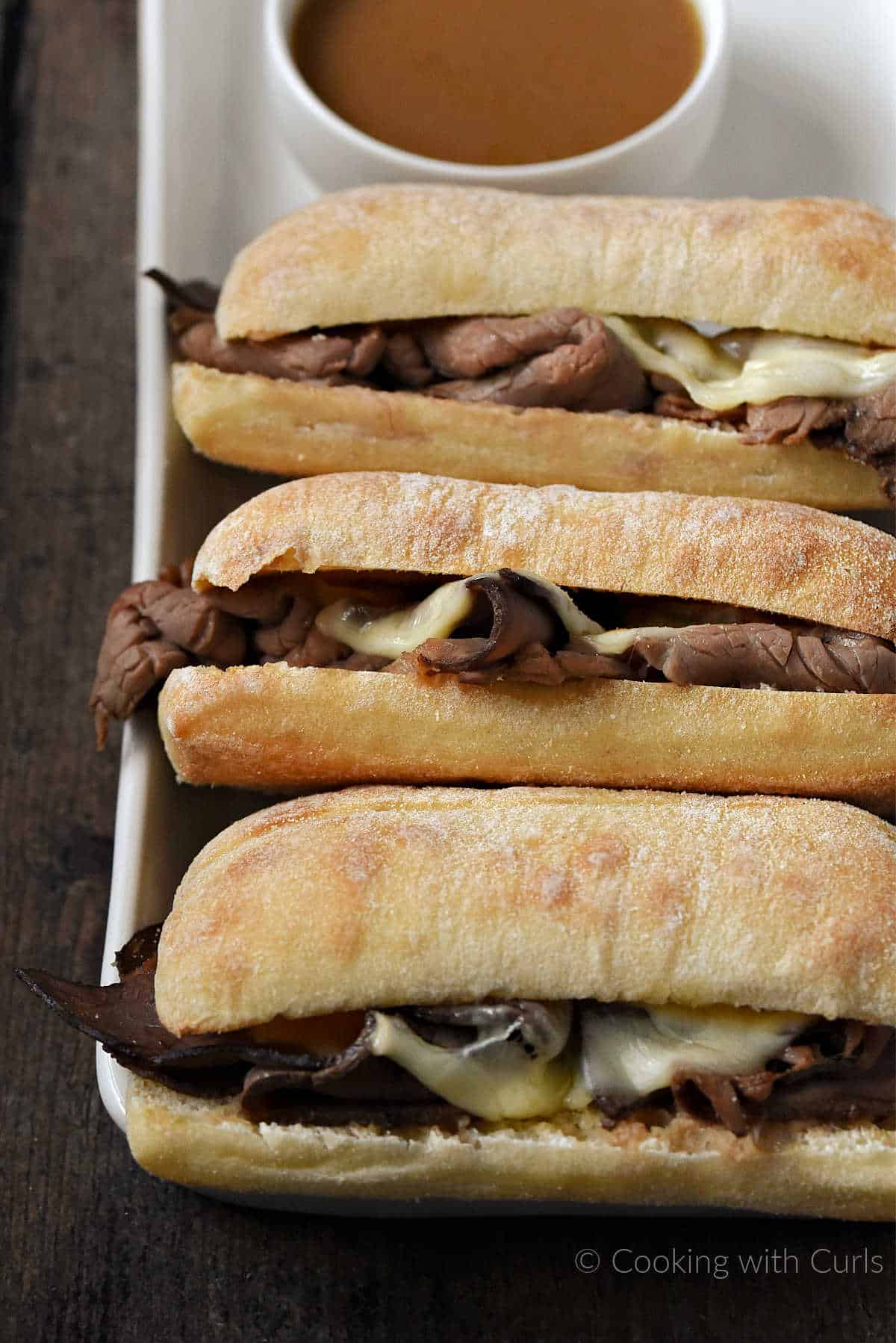 Three French Dip Sandwiches on a platter with a small bowl of au jus. 