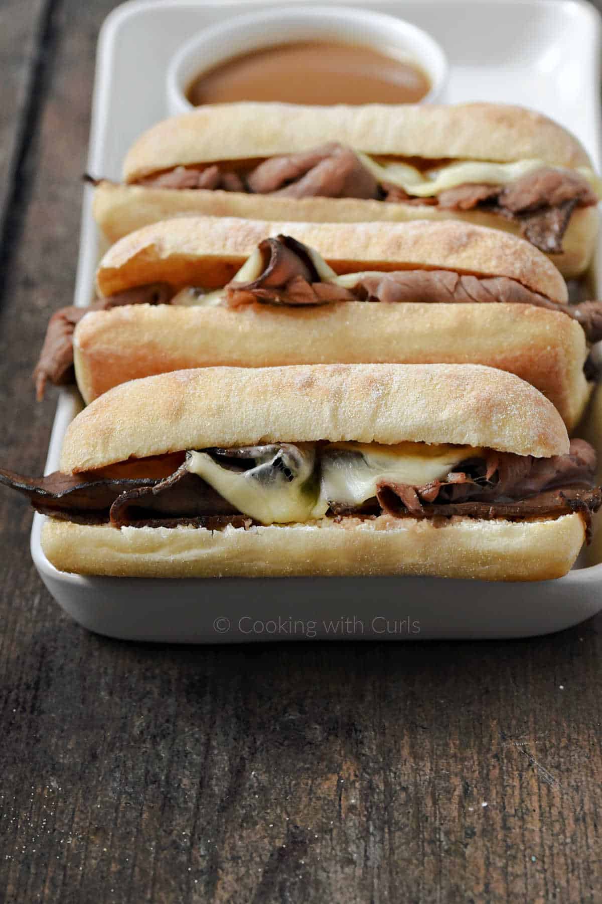 Three French Dip sandwiches on a serving platter with a small bowl of au jus. 