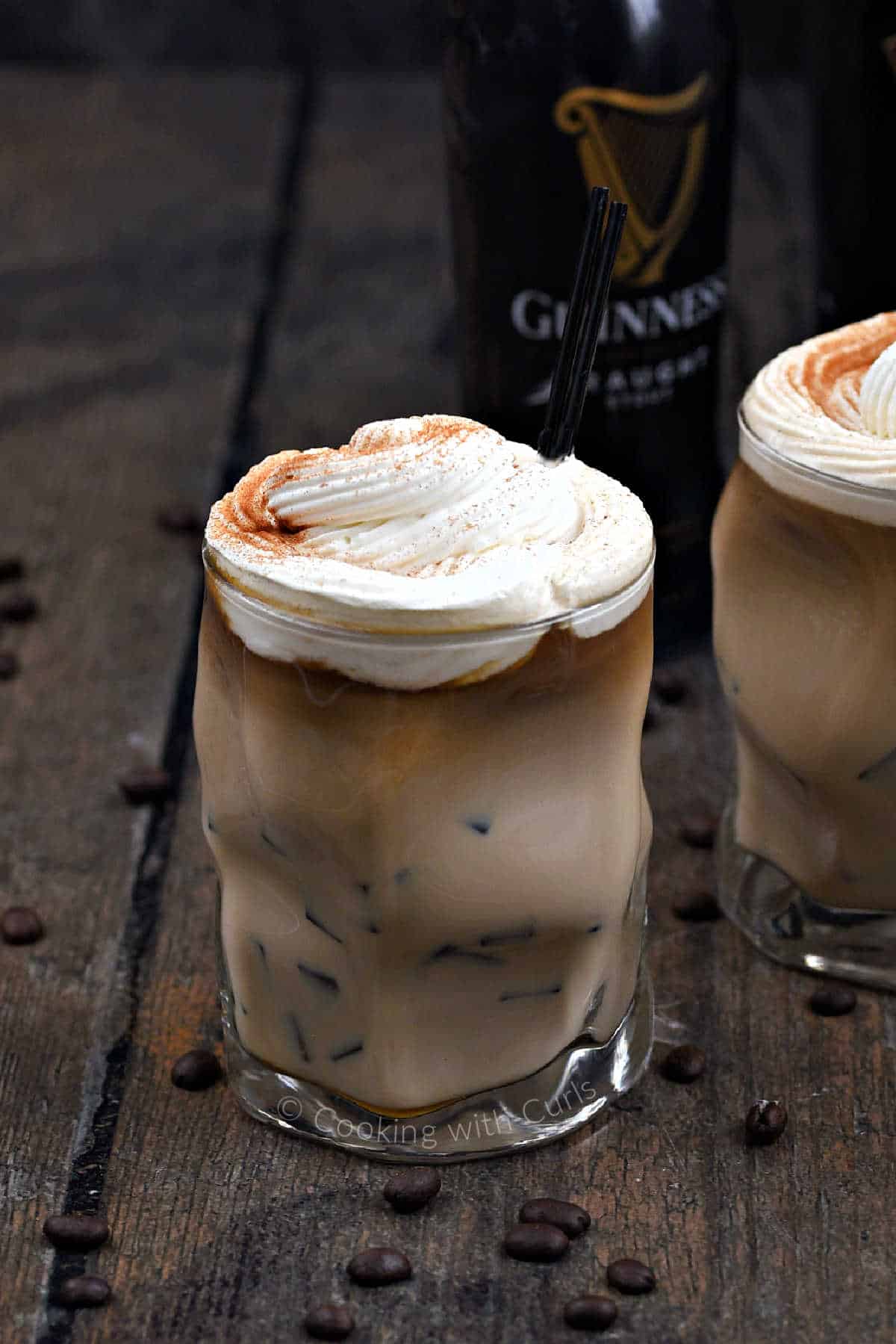Two glasses filled with Irish Iced Coffee topped with whipped cream with a bottle of Guinness in the background.