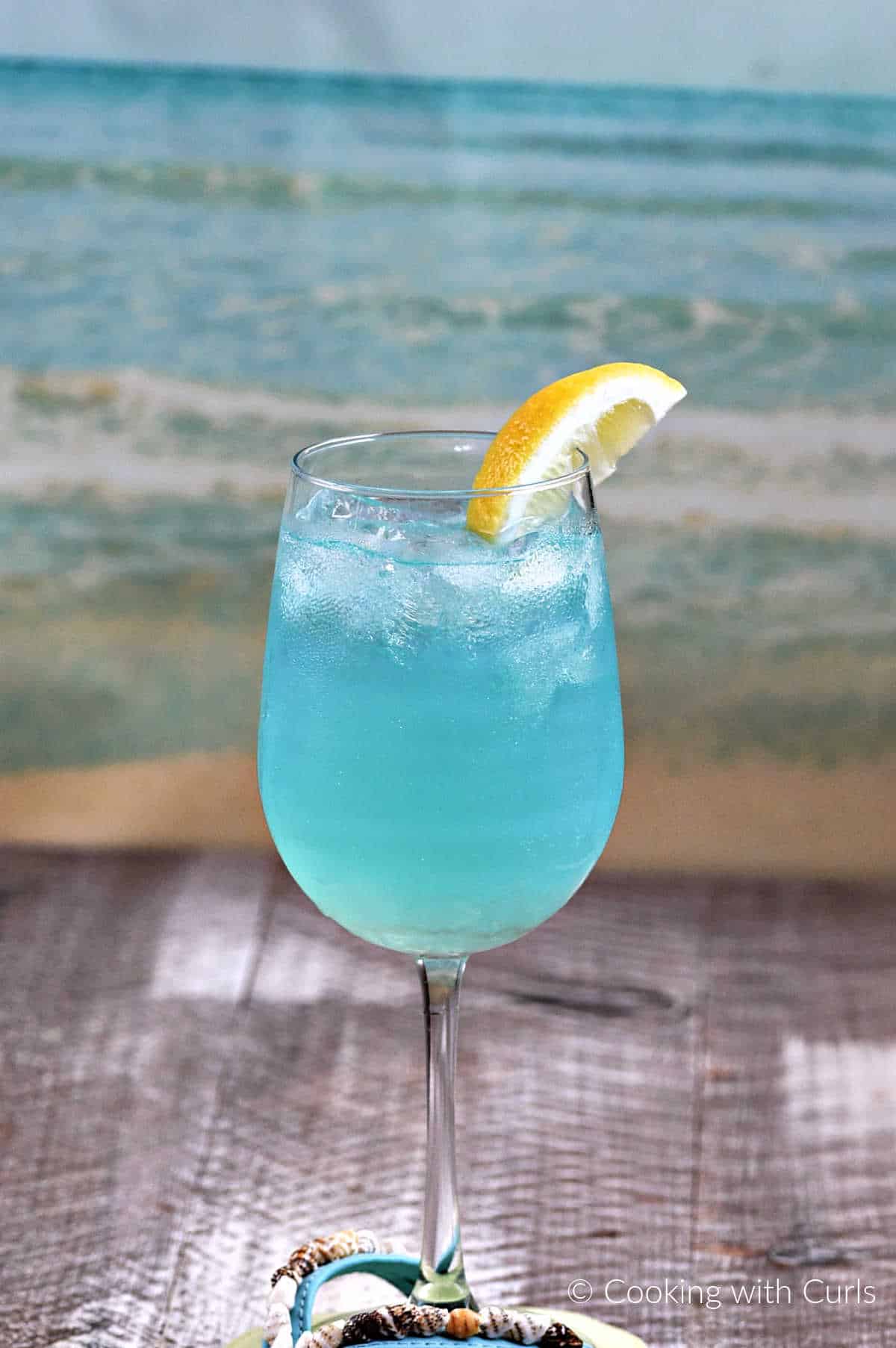Teal blue cocktail with ice in a wine glass with a lemon wedge in front of an ocean waves background.
