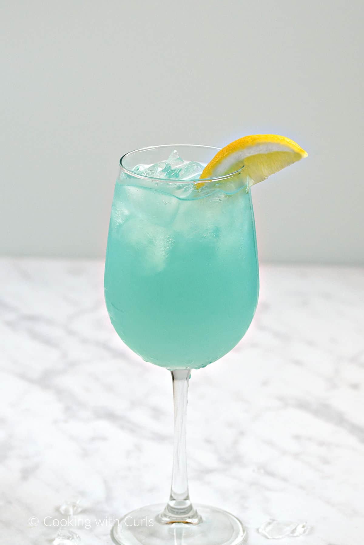 Teal blue cocktail with ice in a white wine glass with a lemon wedge garnish.