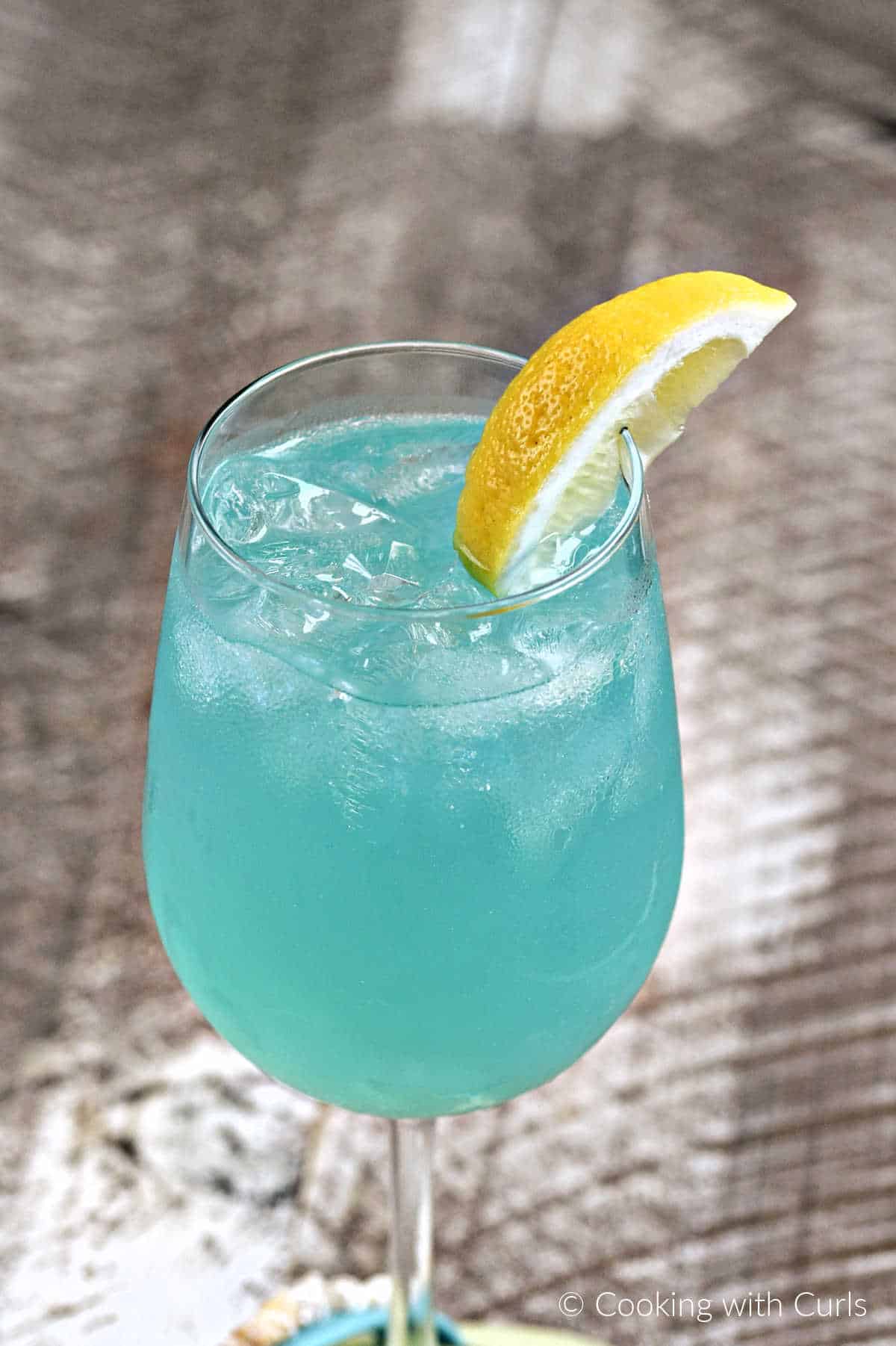 Teal blue cocktail with ice cubes in a wine glass with a lemon wedge on the rim.