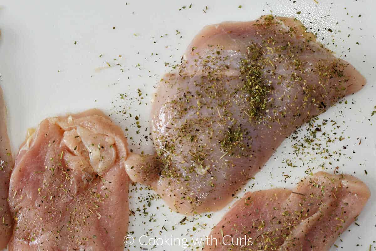 Four chicken breasts sprinkled with Italian seasoning on a cutting board.