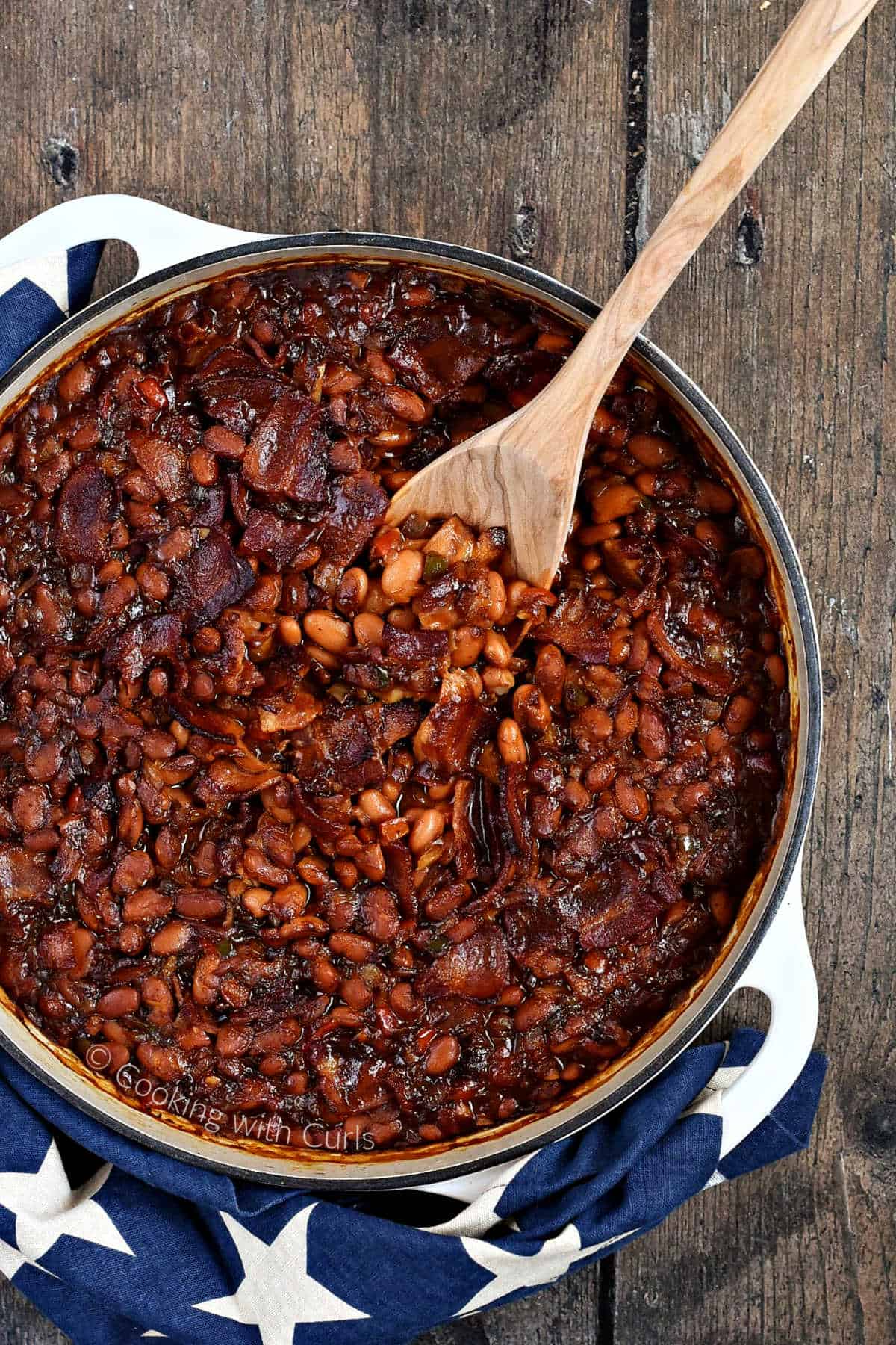 Looking down on a skillet of baked beans with bacon wrapped in a dark blue napkin with white stars and a wooden spoon.
