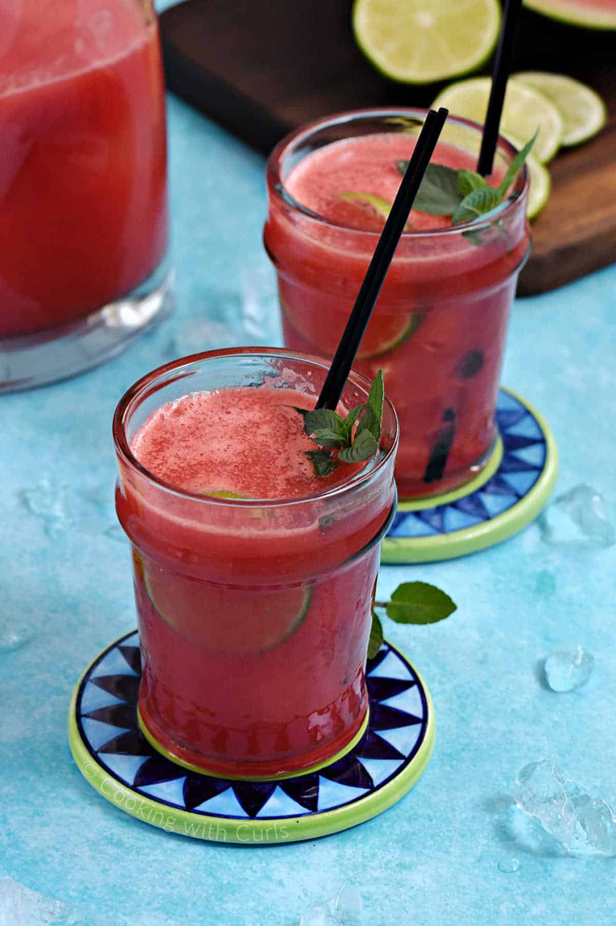 Two glasses of watermelon agua fresca with mint leaves, lime wheels, and cocktail straws, with a pitcher in the background.