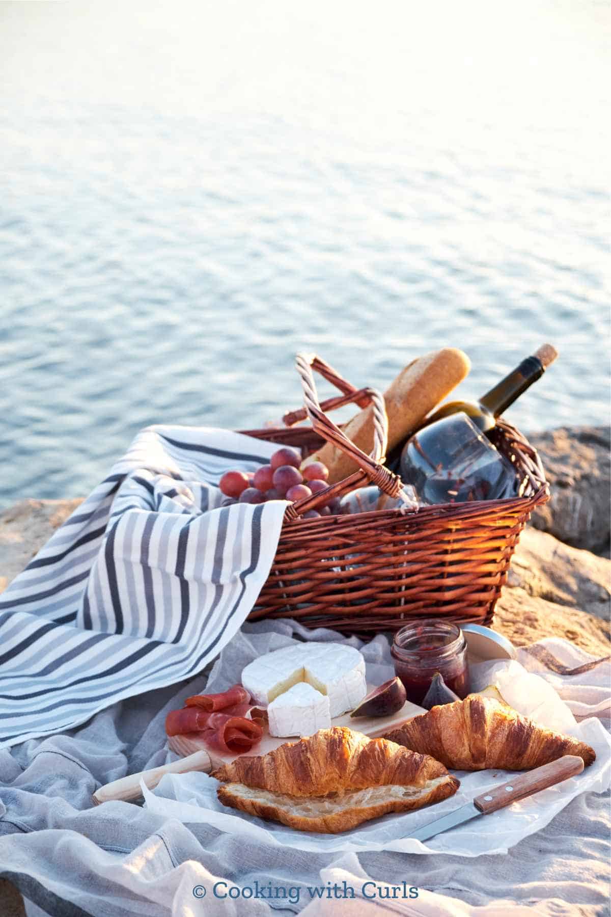 A picnic basket filled with grapes, wine, and baguette sitting on a blanket with croissants, brie, and jam near the water.