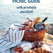 A picnic basket filled with grapes, wine, and baguette sitting on a blanket with croissants, brie, and jam near the water with title graphic across the top.