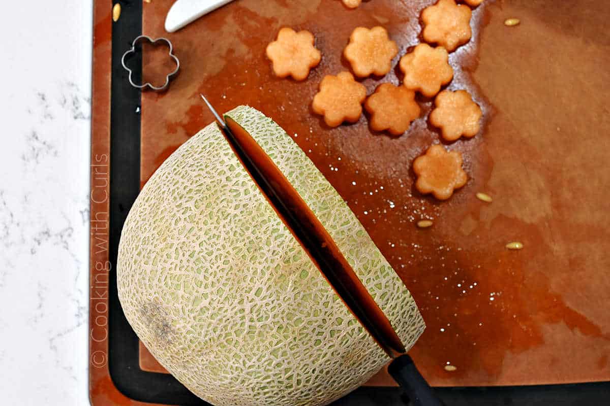 A knife slicing through the side of a cantaloupe with flower shaped cut-outs in the background.