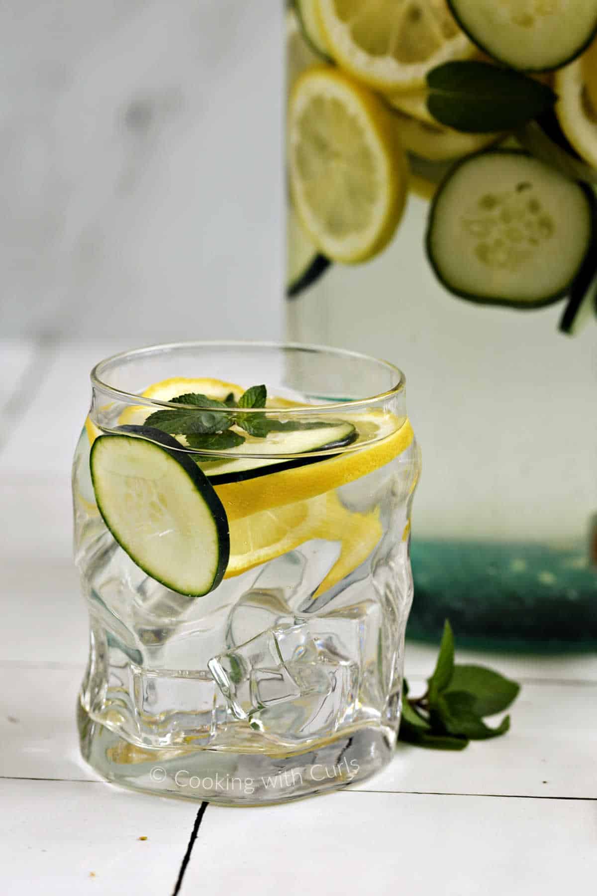 An ice filled glass of cucumber, lemon, and mint infused water with a large jar of infused water in the background.