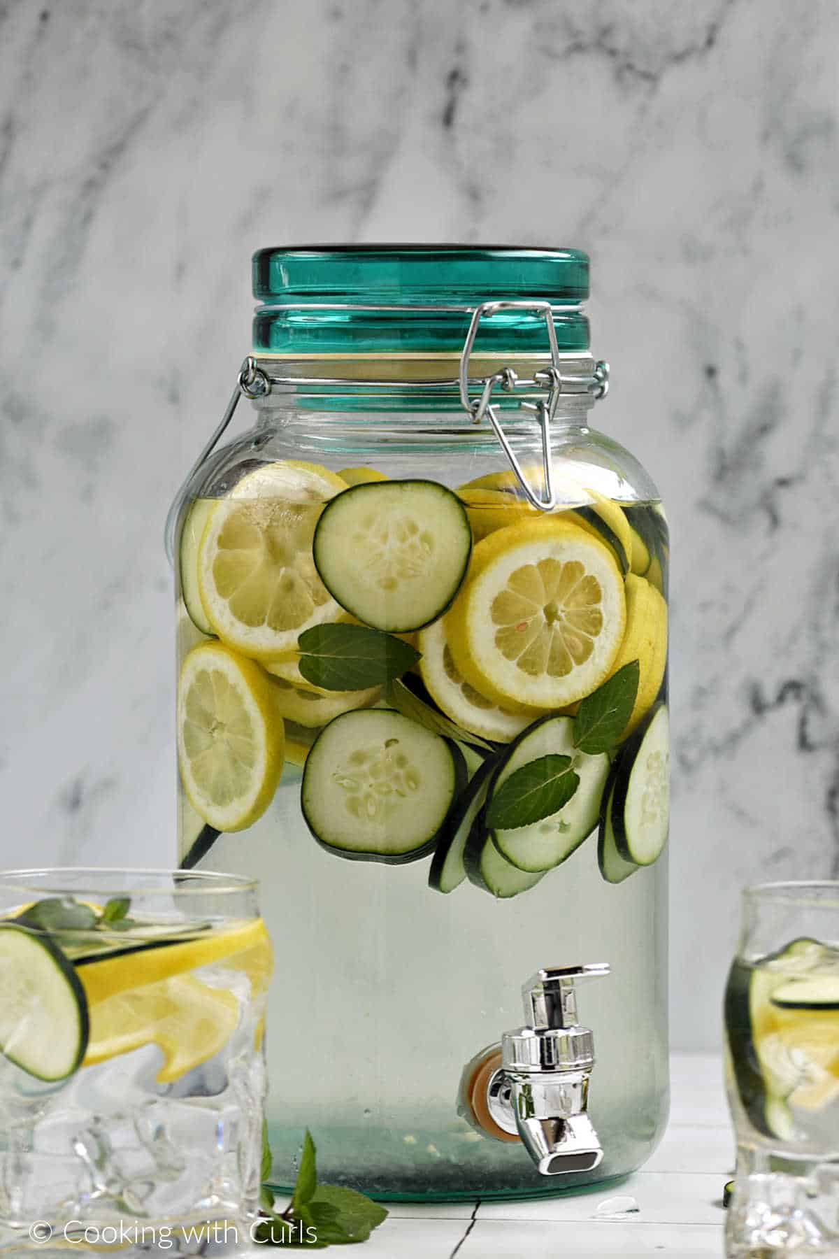A gallon canister filled with lemon and cucumber slices, mint leaves, and water.