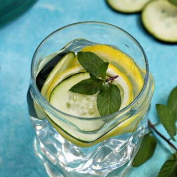 A glass of cucumber lemon mint water with slices of cucumber and sprigs of mint in the background.
