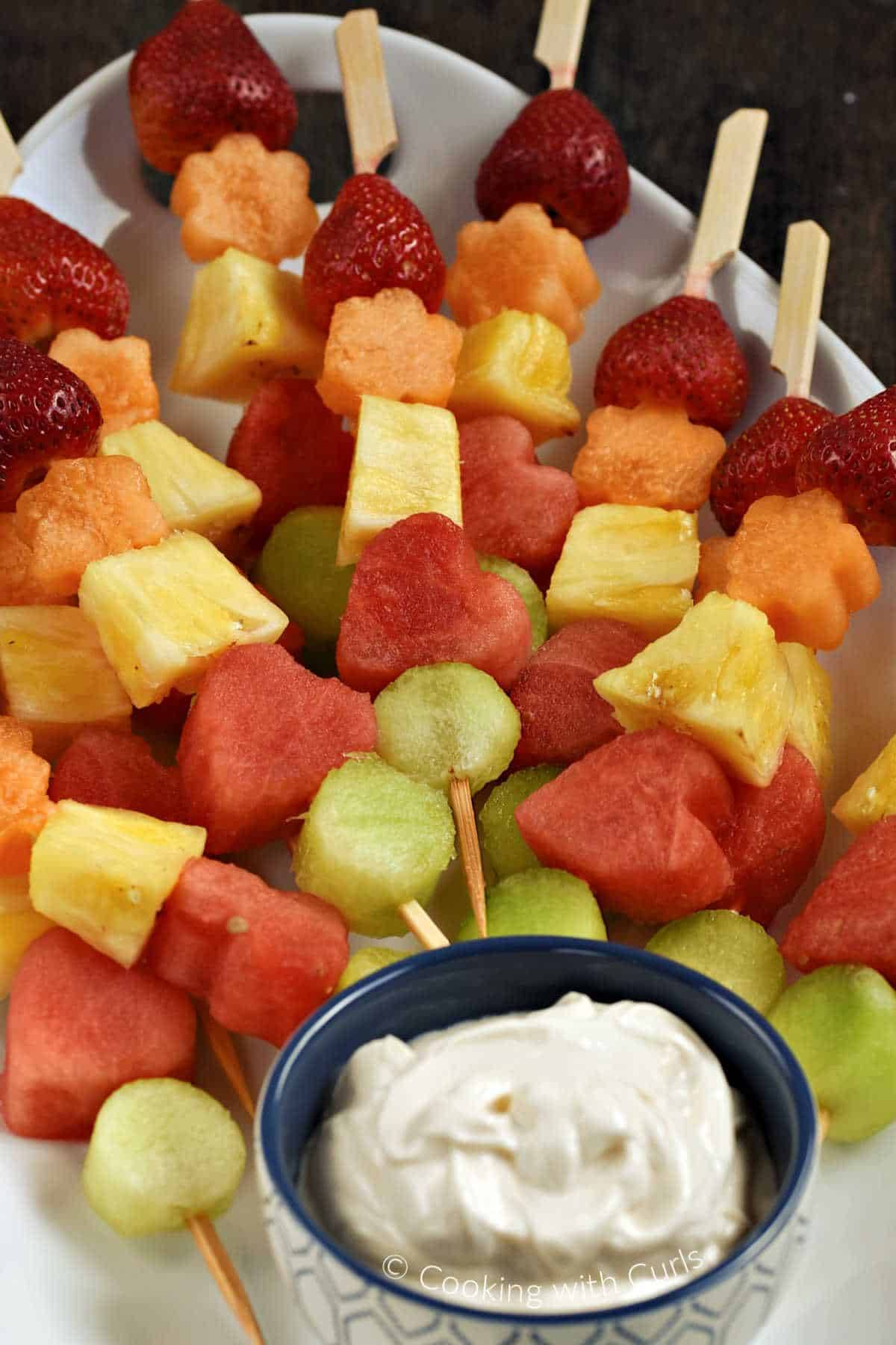 Close-up image of fresh fruits threaded onto wood skewers, stacked on an oval platter with a bowl of yogurt dip at the bottom.