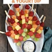 Fresh fruits threaded onto wood skewers, stacked on an oval platter with a bowl of yogurt dip at the bottom and title graphic across the top.